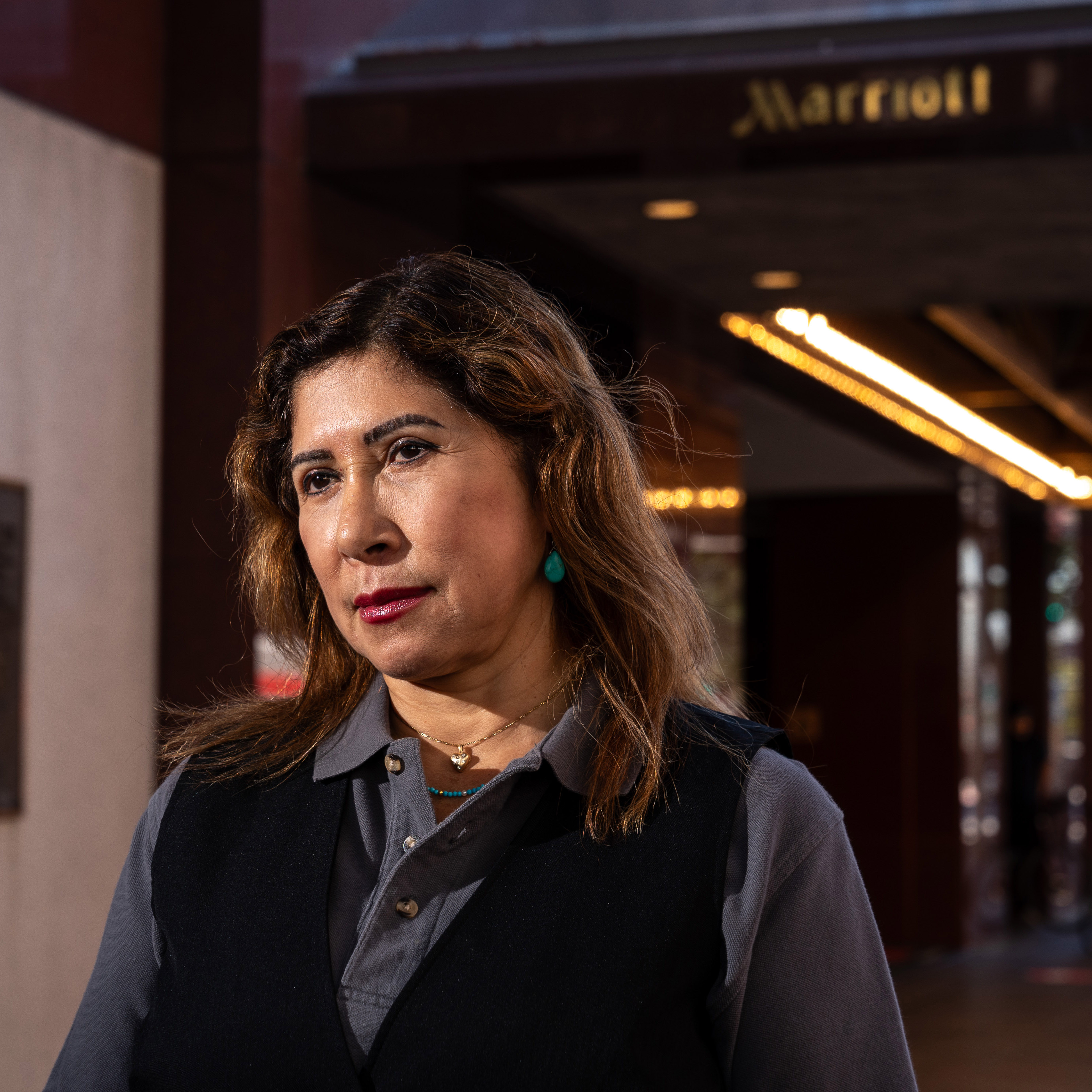 A woman with wavy hair is standing outside, wearing a gray blouse with a black vest, under an illuminated canopy with a sign that reads "Marriott" in the background.