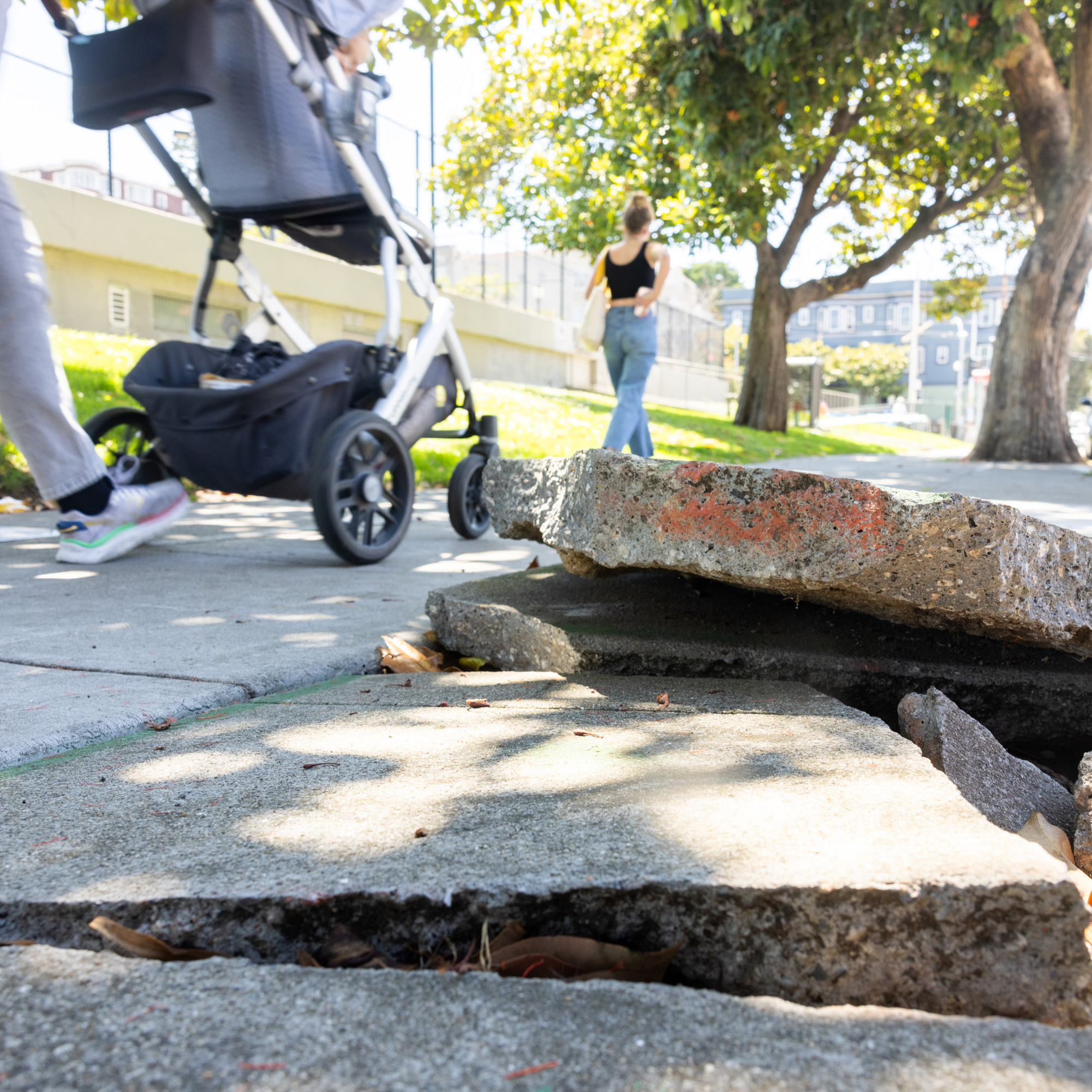 A sidewalk with large, cracked, and uneven concrete slabs is shown. A blurred motion of a person pushing a stroller and another person walking are in the background.