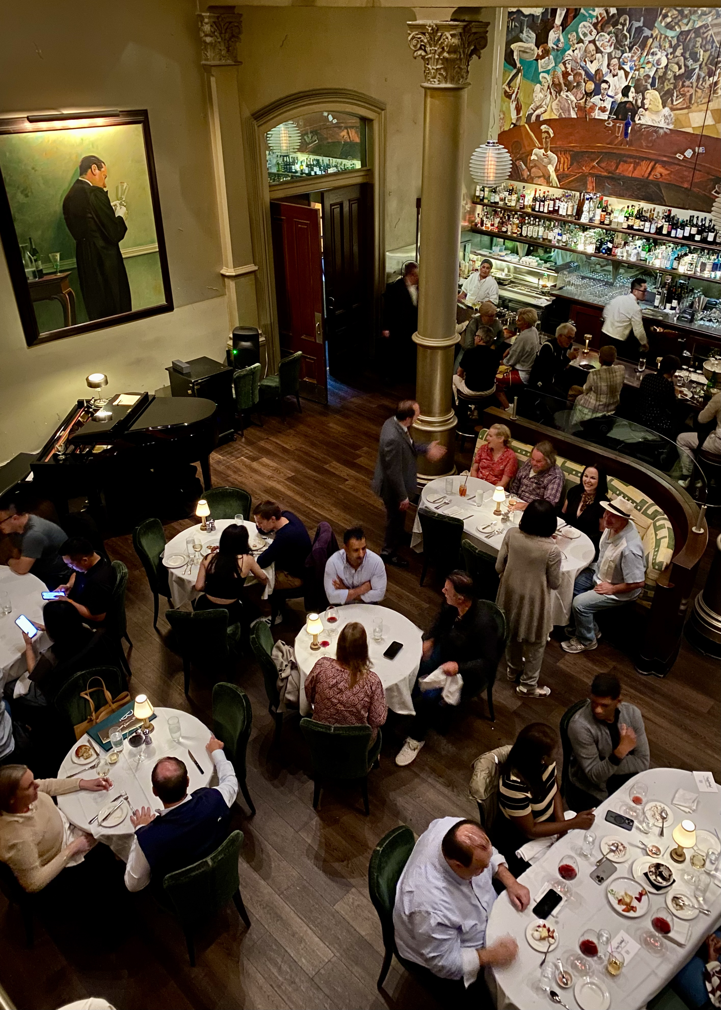 A lively restaurant scene with patrons seated at round tables, a bar staffed by bartenders, and framed paintings on the walls, including one large one of a person in profile.