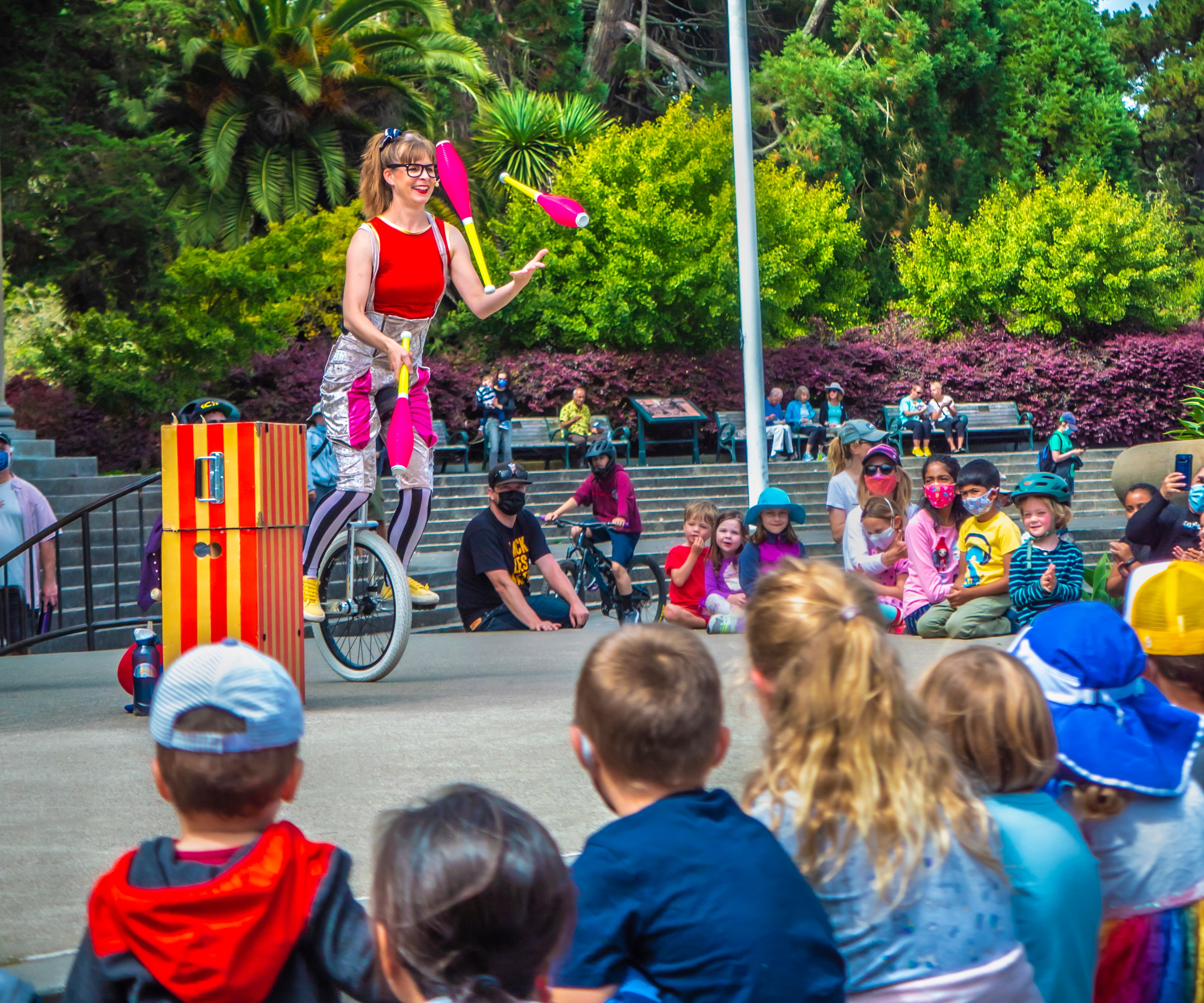 A performer juggles clubs while riding a unicycle before an audience of children and adults in a park, with lush greenery in the background.