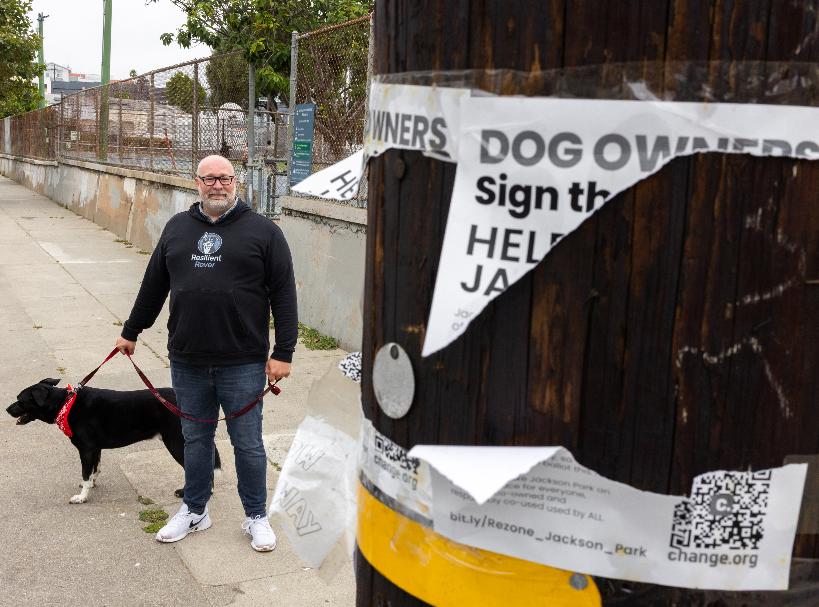 A man in a black &quot;Resilient Rover&quot; hoodie walks a black dog bandana in an urban setting near a utility pole with torn dog owner posters calling for signatures.