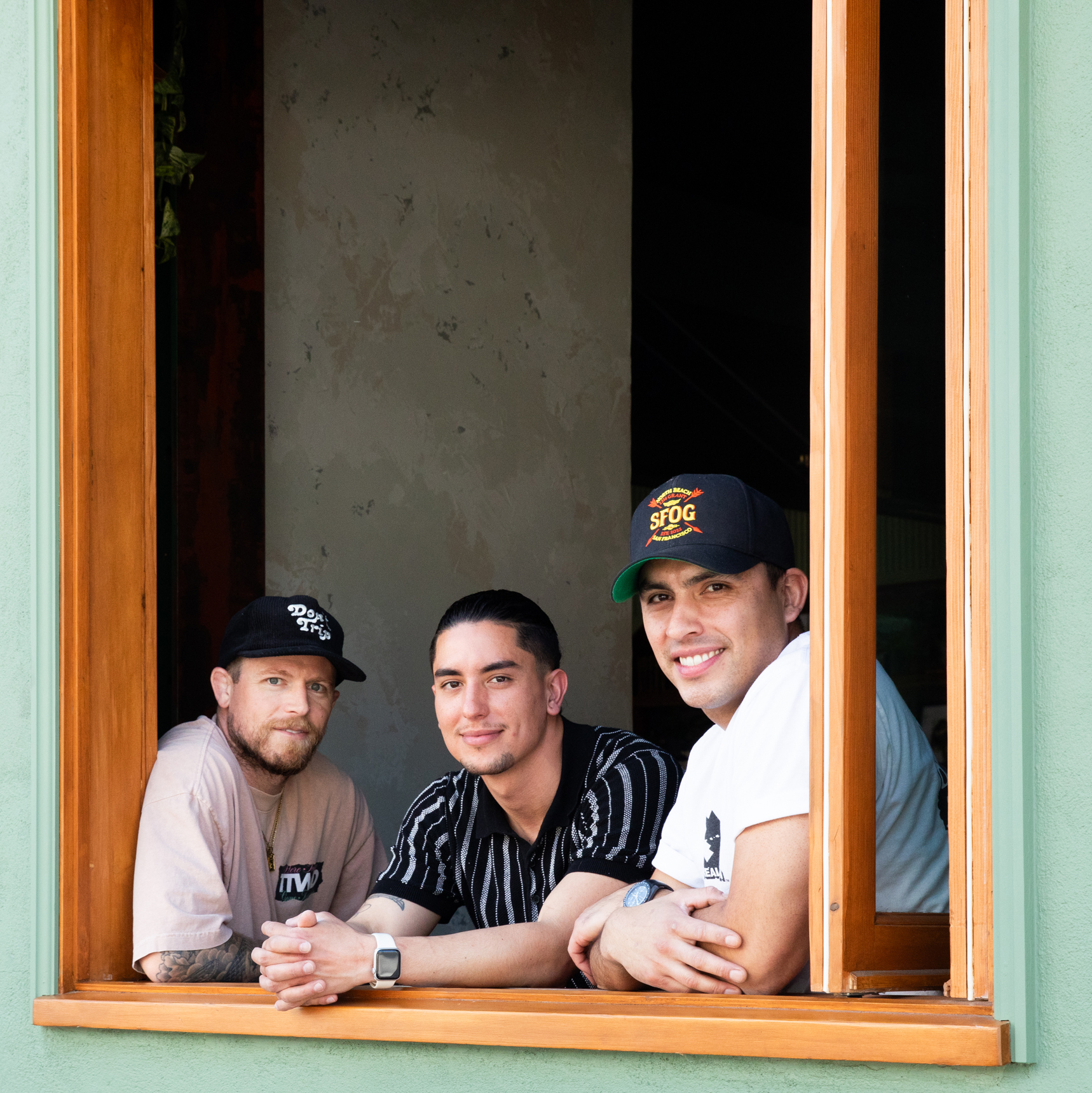 Three men wearing casual clothing and hats lean on a wooden window frame, smiling and looking at the camera, with a muted, indoor background behind them.