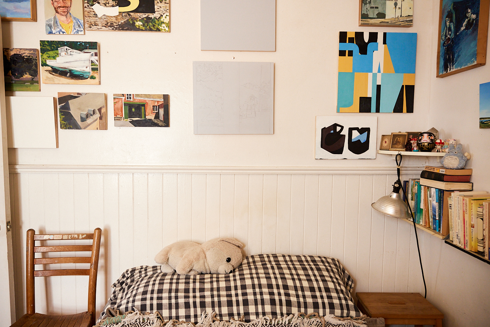 The image shows a cozy room with a wooden chair, a bed with a checkered blanket and stuffed bear, bookshelves, framed artwork, and a desk lamp on the wall.