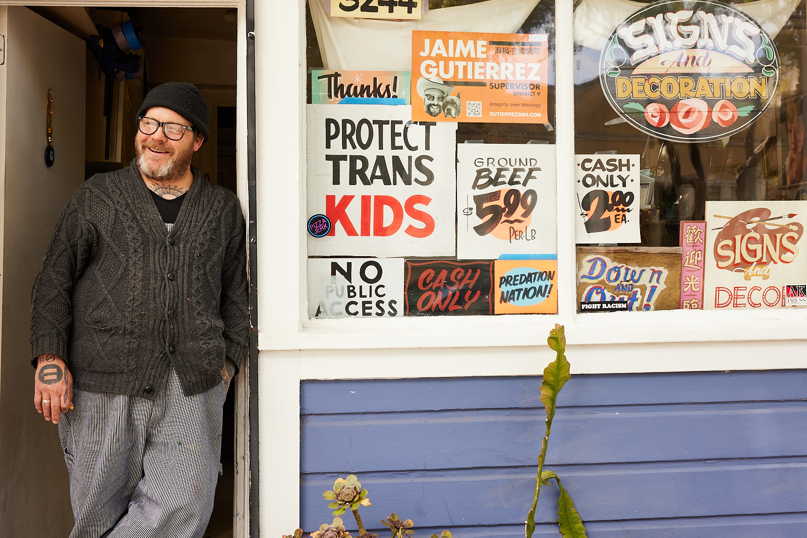 A person stands smiling at the doorway of a shop with various signs in the window, including "Protect Trans Kids," "Ground Beef $5.99 per lb," and "Cash Only $2.00 ea."
