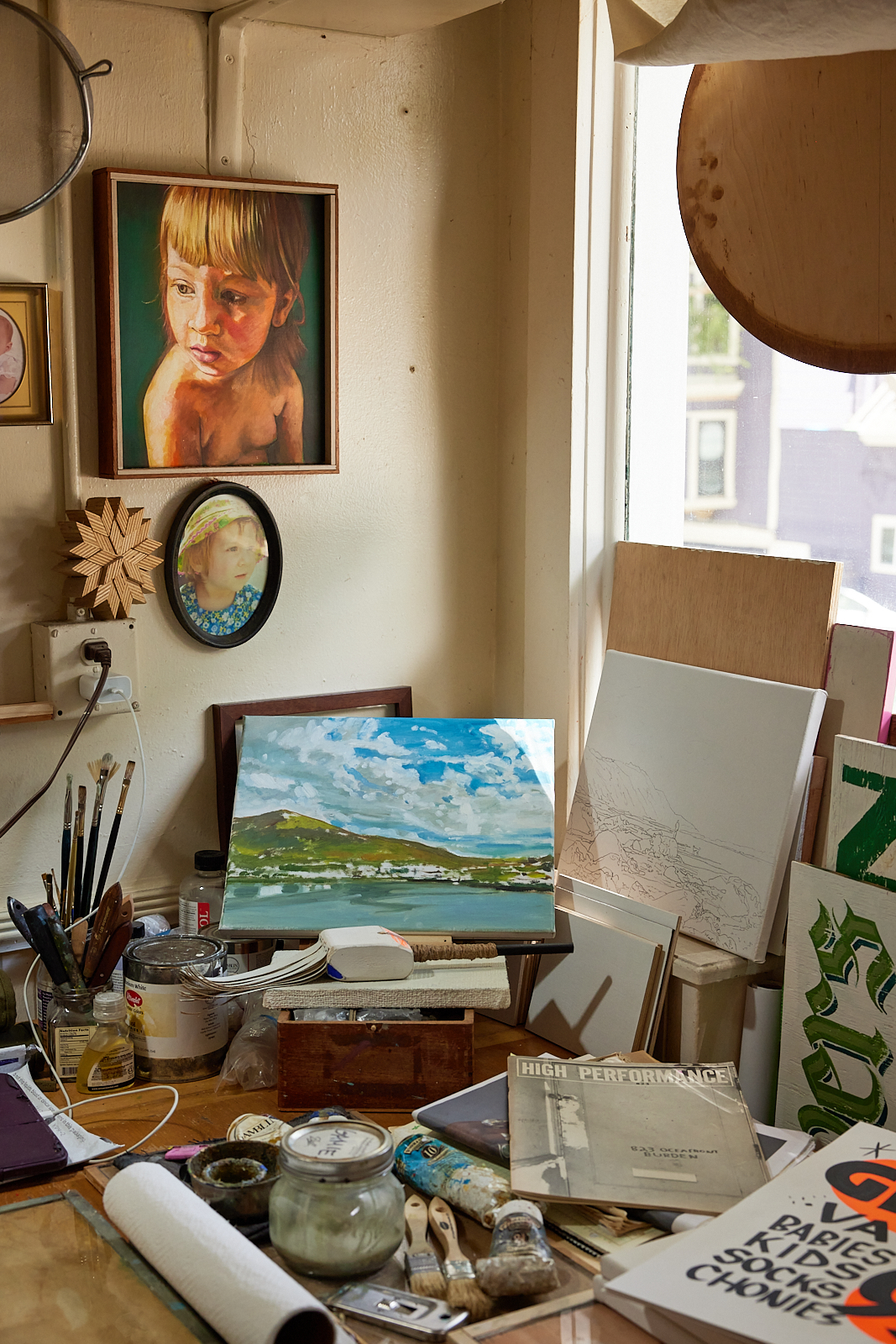 The image shows an artist's studio with paintings on the wall, a landscape painting on an easel, various brushes, jars, and art supplies scattered on a cluttered wooden table.