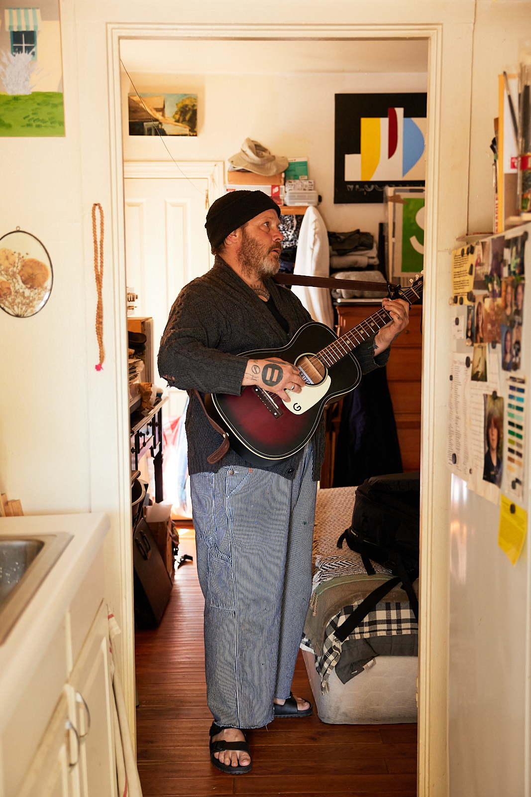 A bearded man in a beanie and casual clothing plays an acoustic guitar in a small, cluttered room with artworks on the wall and various belongings around him.