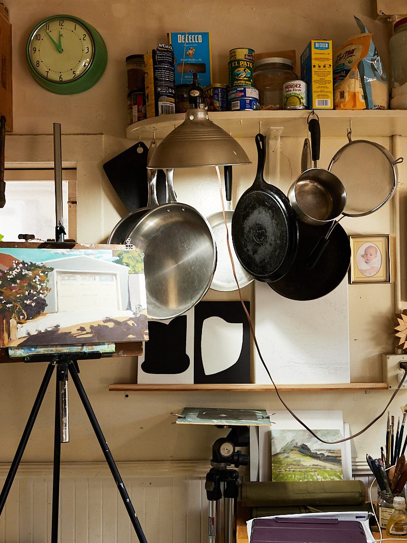 The image depicts an artist's studio with paintings, an easel, and paint supplies. Pots and pans hang from the wall, and shelves are cluttered with jars and various items.