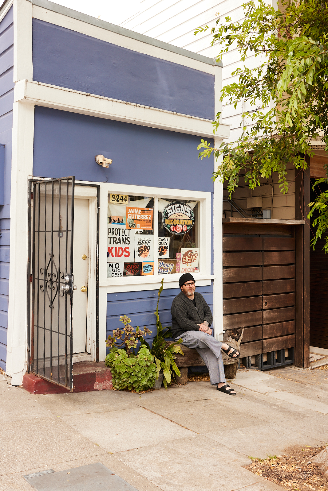 A man sits on a bench outside a blue storefront at 3244. The window is adorned with various colorful signs, and a metal gate is slightly open beside plants.