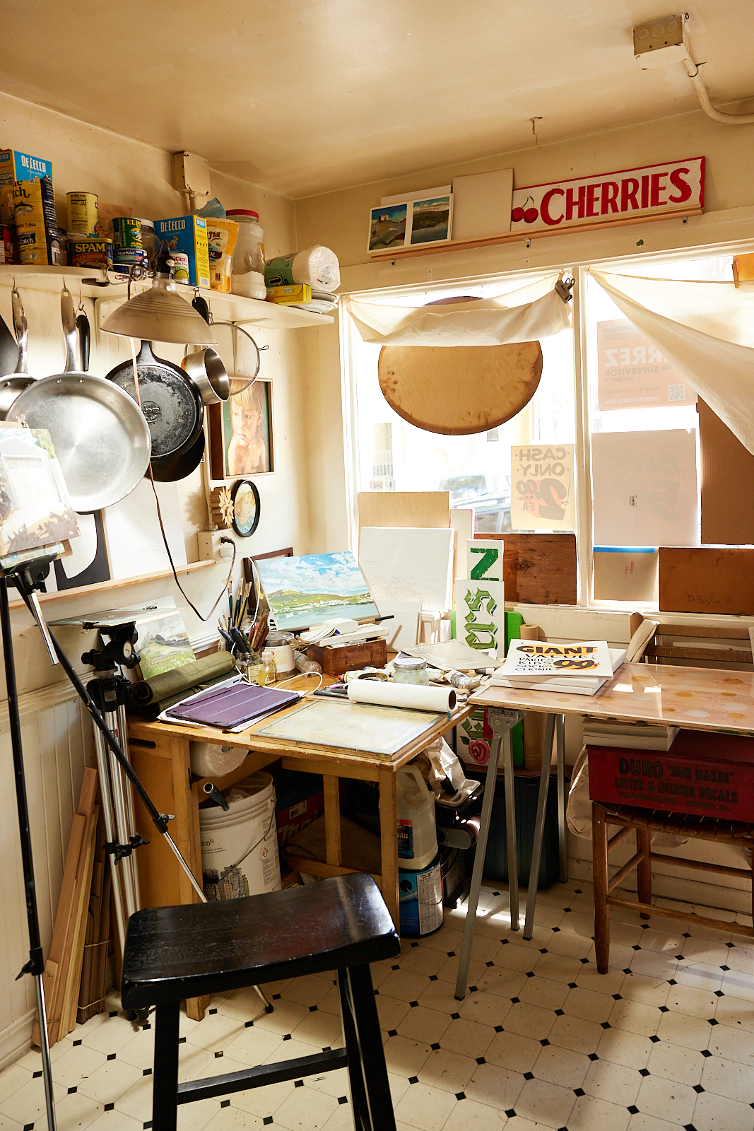 This image shows a cluttered art studio with various supplies. Shelves hold kitchen items, while paintings, brushes, and signs fill the room, lit by a soft, natural light.