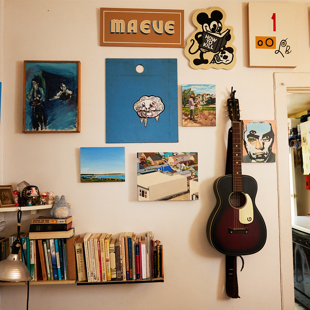 A wall with various paintings, a guitar, and a bookshelf containing books and small ornaments, including a lamp, creating a cozy and artistic ambiance.