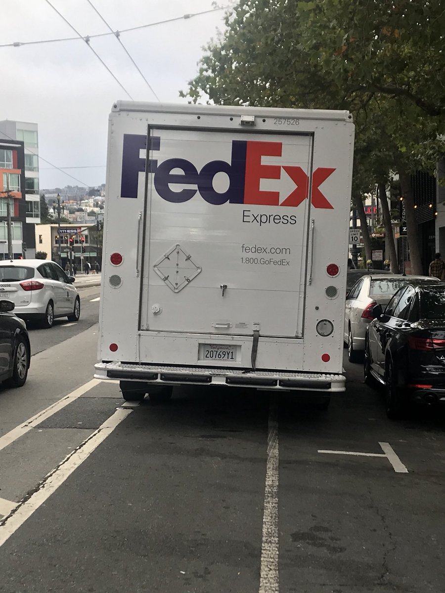 A white FedEx truck is parked in a bike lane on a city street.