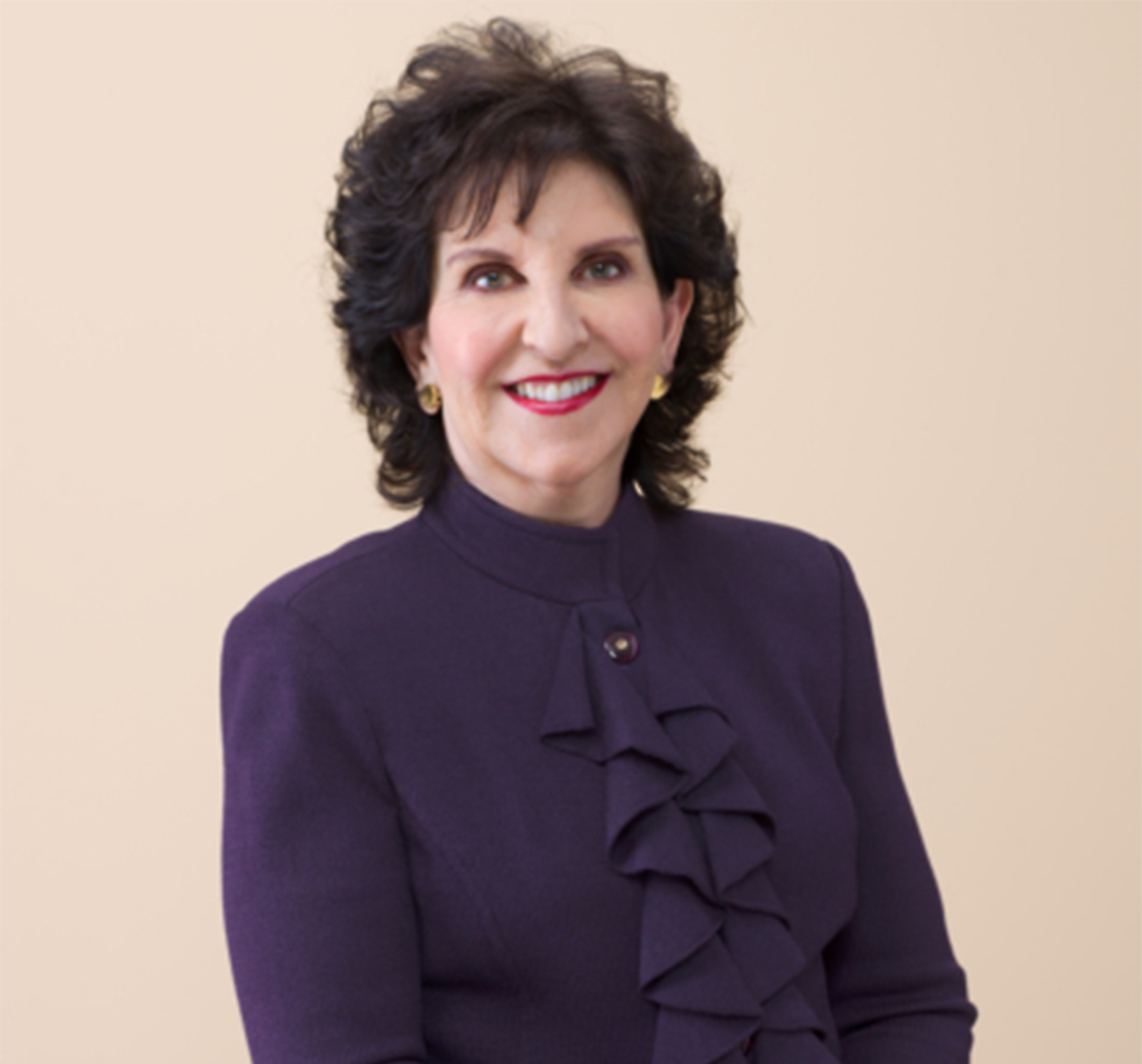 A woman with dark, curly hair and gold earrings smiles. She's wearing a purple top with ruffled detailing on the front and stands against a beige background.