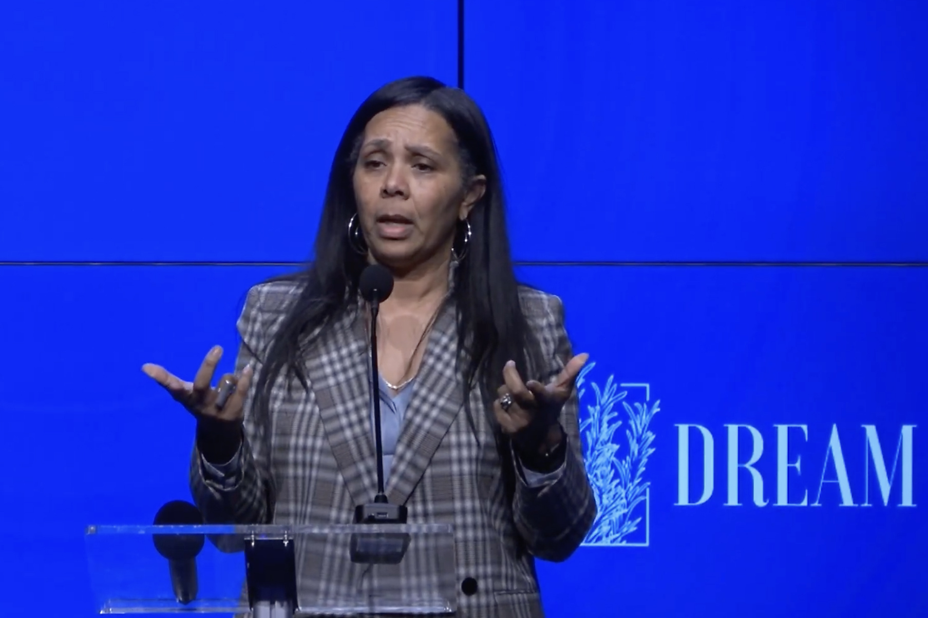 A woman with long hair speaks into a microphone. She is wearing a plaid blazer with a blue shirt and gesturing with both hands. The background is a blue screen with &quot;DREAM&quot; text.