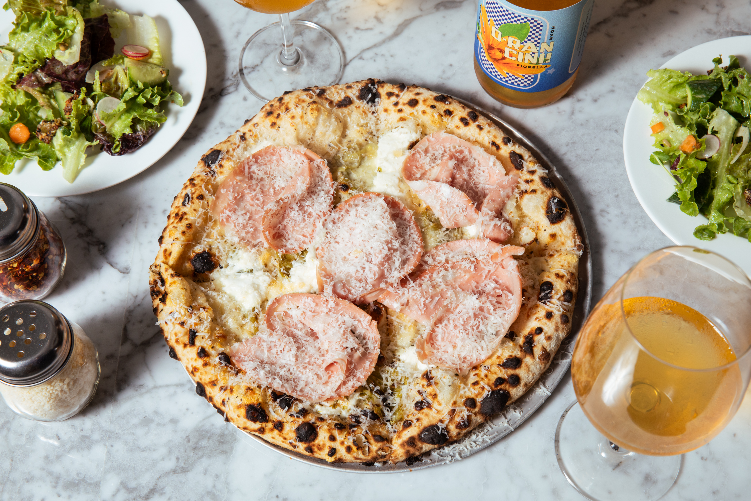 A pizza topped with cheese and slices of meat is on a marble table, accompanied by two plates of salad, a glass of beer, a beer bottle, and seasoning shakers.