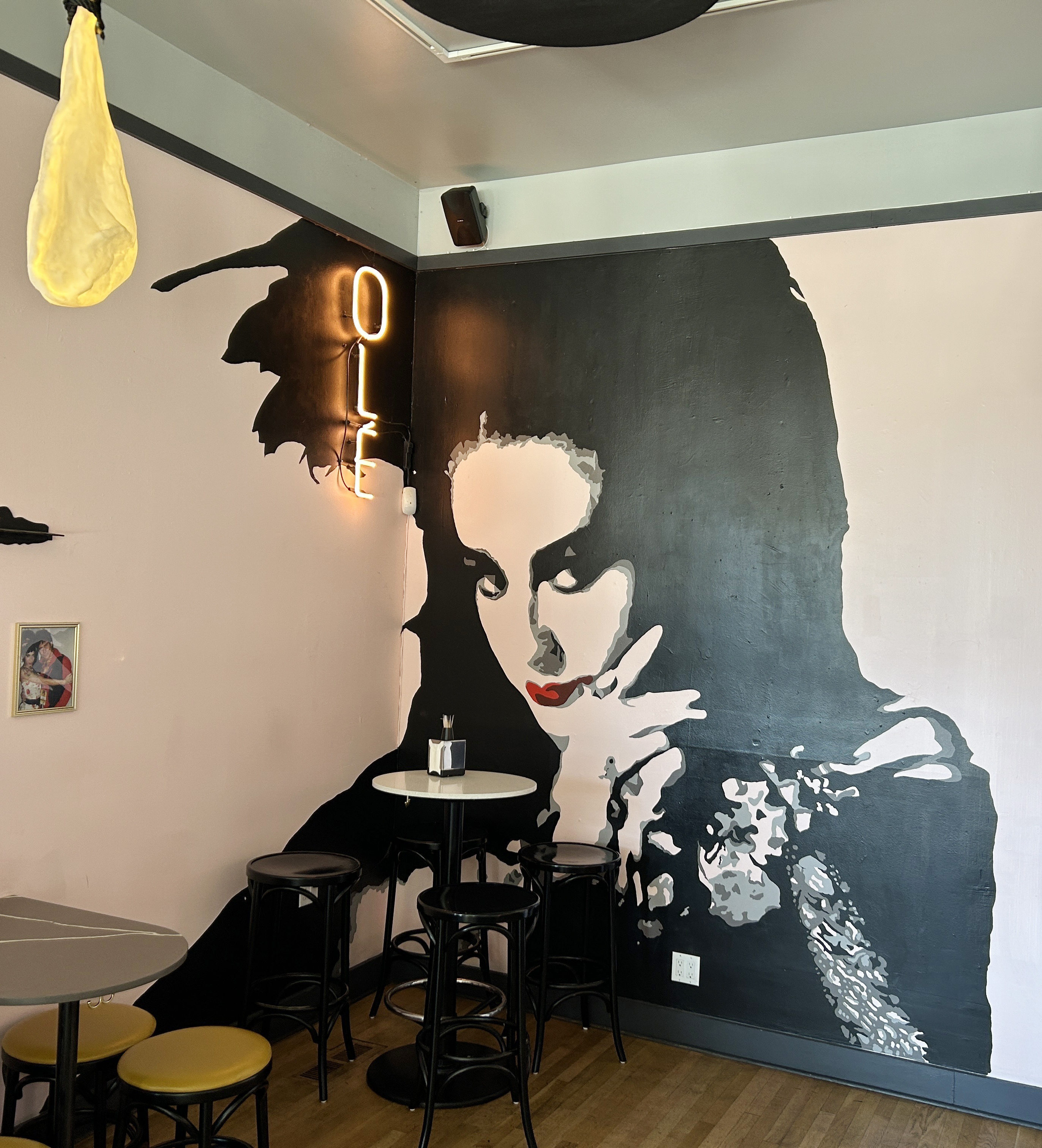 A small cafe corner has high stools and tables, a large painted mural of a pensive woman with bright red lips, and a neon &quot;OLE&quot; sign on the wall.