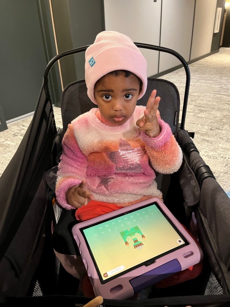 A young child wearing a pink beanie and a colorful sweater is sitting in a stroller, holding one hand up. An iPad with a kids' app is on their lap.