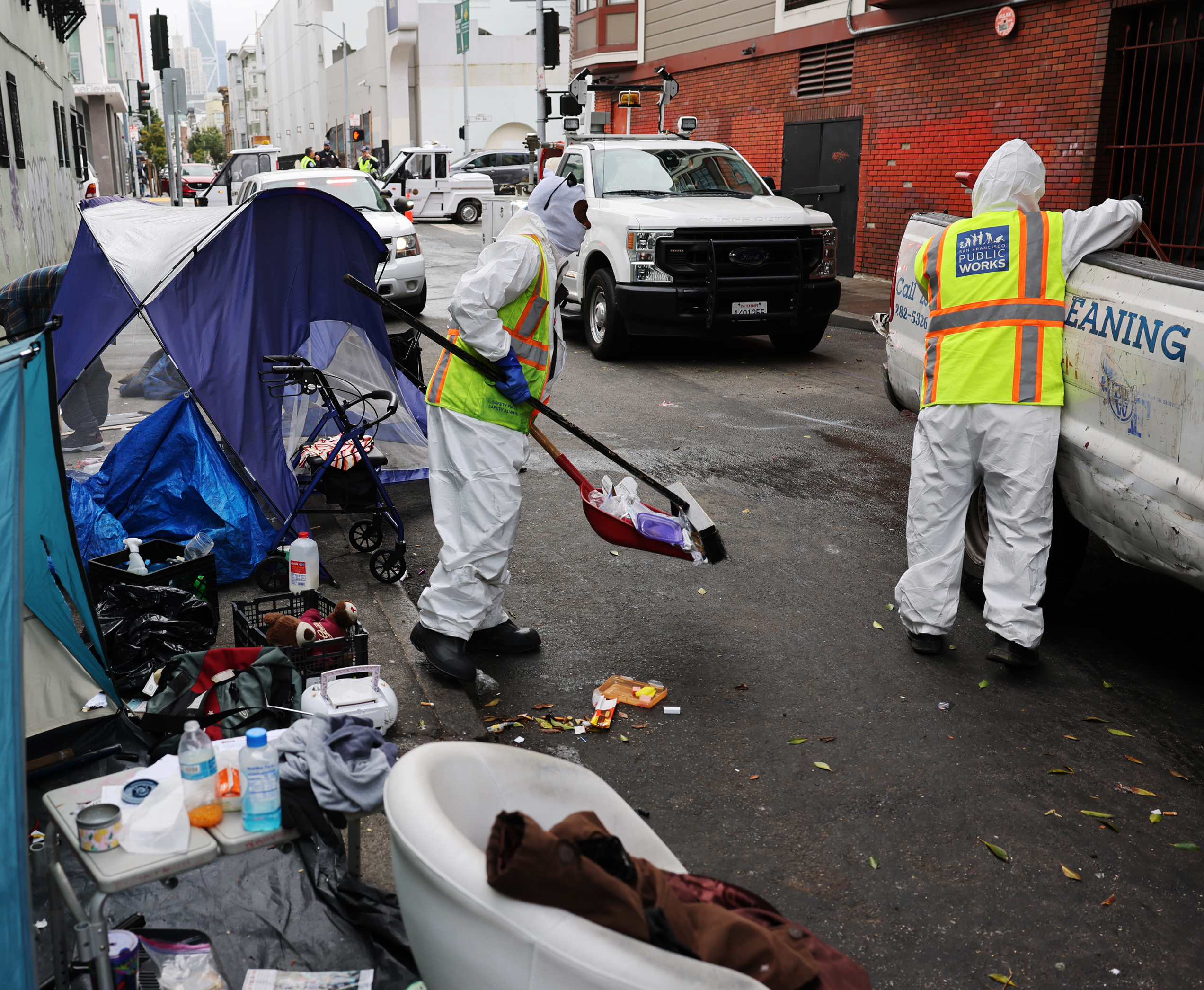 Workers in protective suits clean a street area cluttered with trash and a tent. One sweeps debris into a dustpan while the other leans against a cleaning truck.