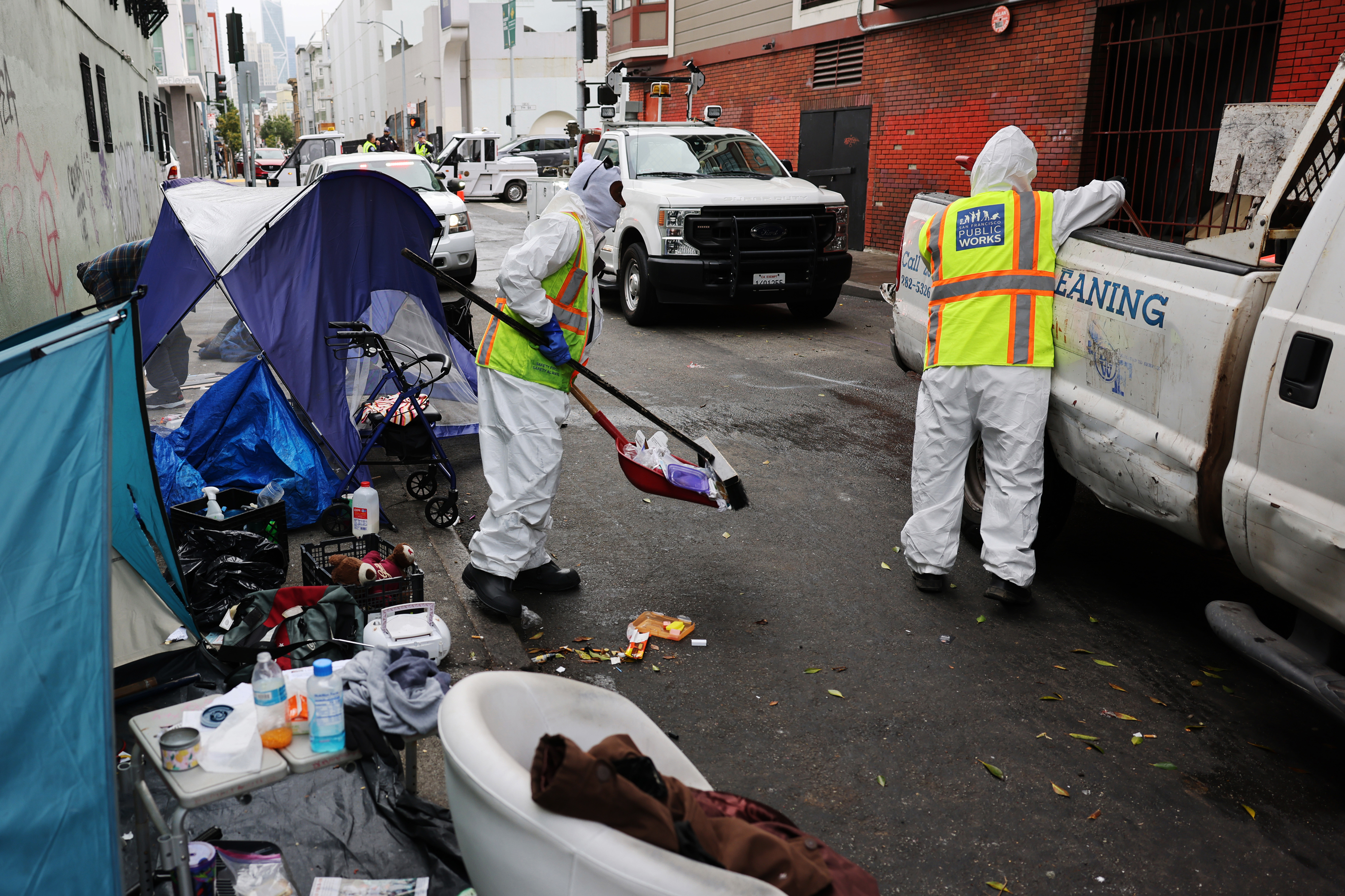 Two workers in hazmat suits and neon vests clean up a cluttered street near a tent. They load trash into a truck as various discarded items are scattered around.
