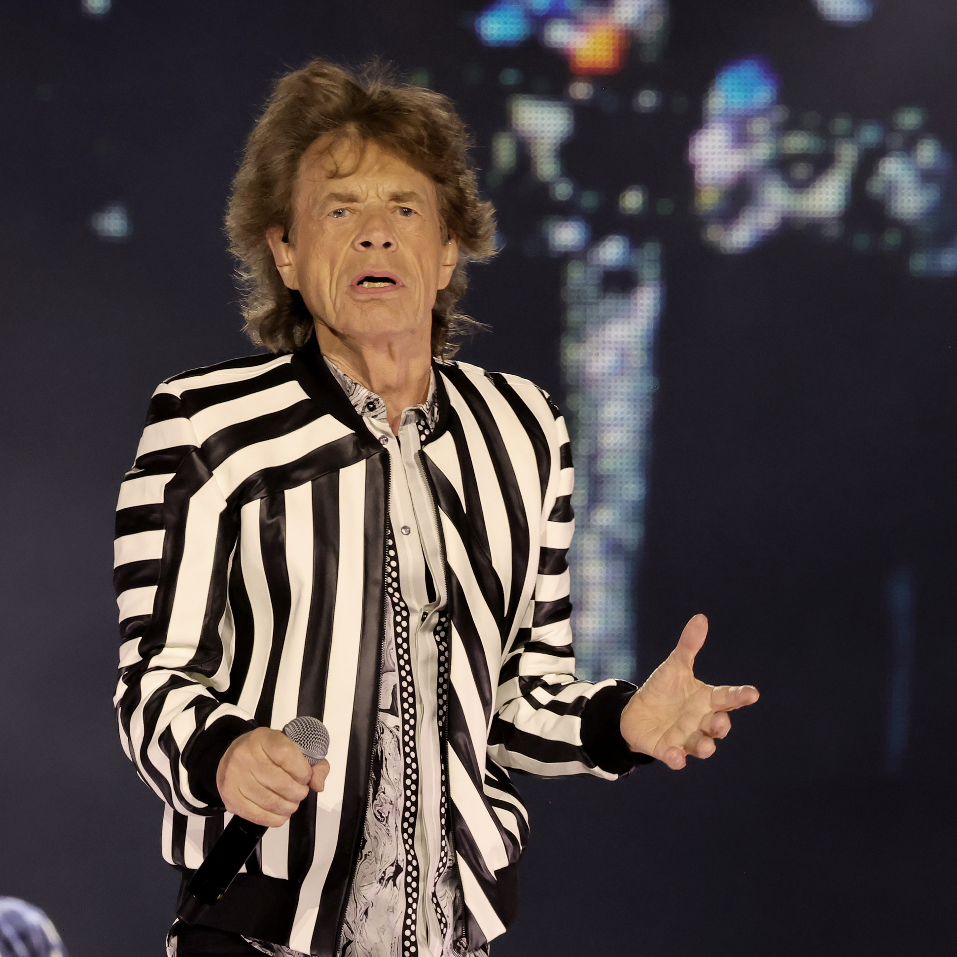 An older man with tousled hair is wearing a black and white striped jacket. He holds a microphone in his right hand and is gesturing with his left hand, performing on stage.