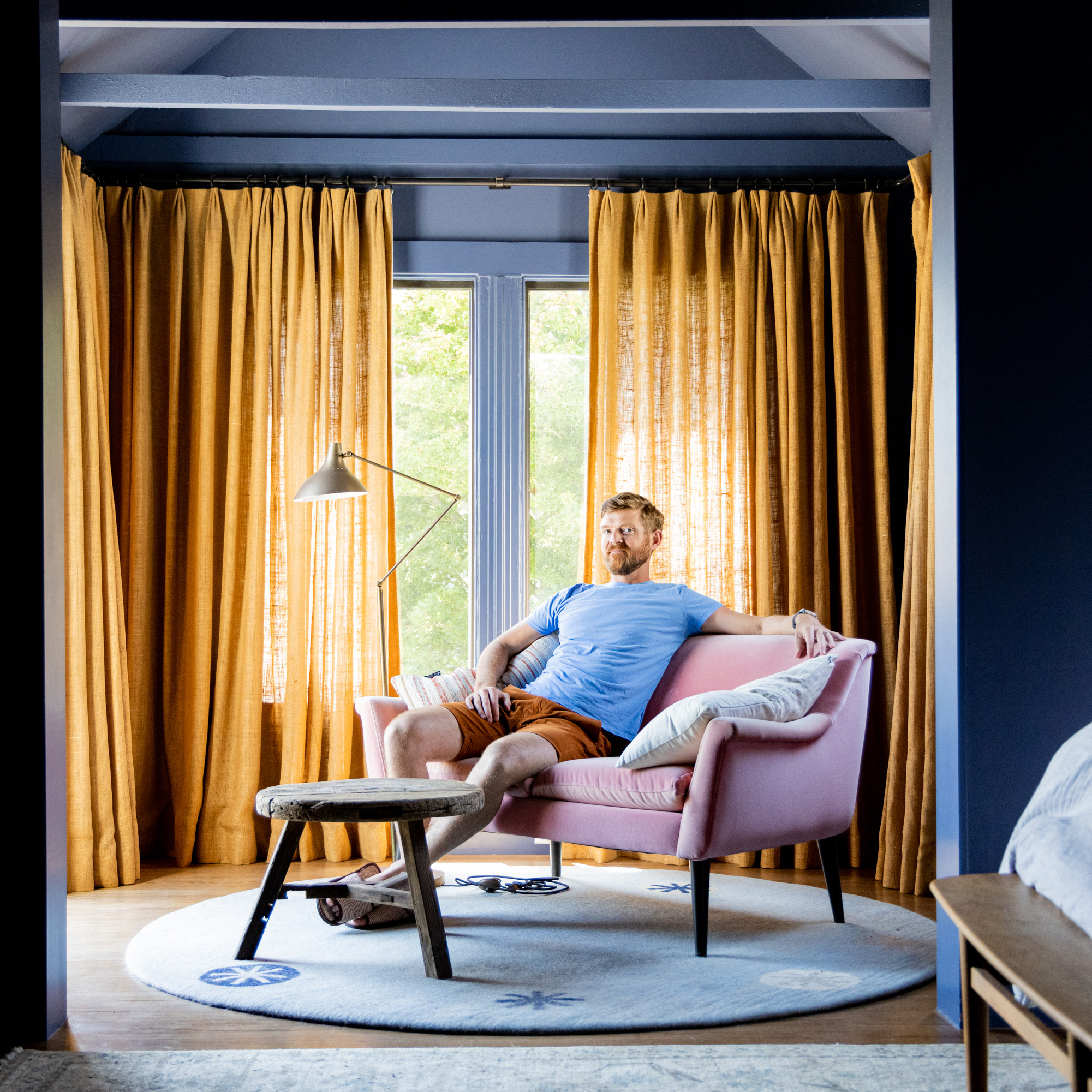 A man in a blue shirt and brown shorts relaxes on a pink sofa in a cozy room with large windows, yellow curtains, a modern floor lamp, and a rustic wooden stool.