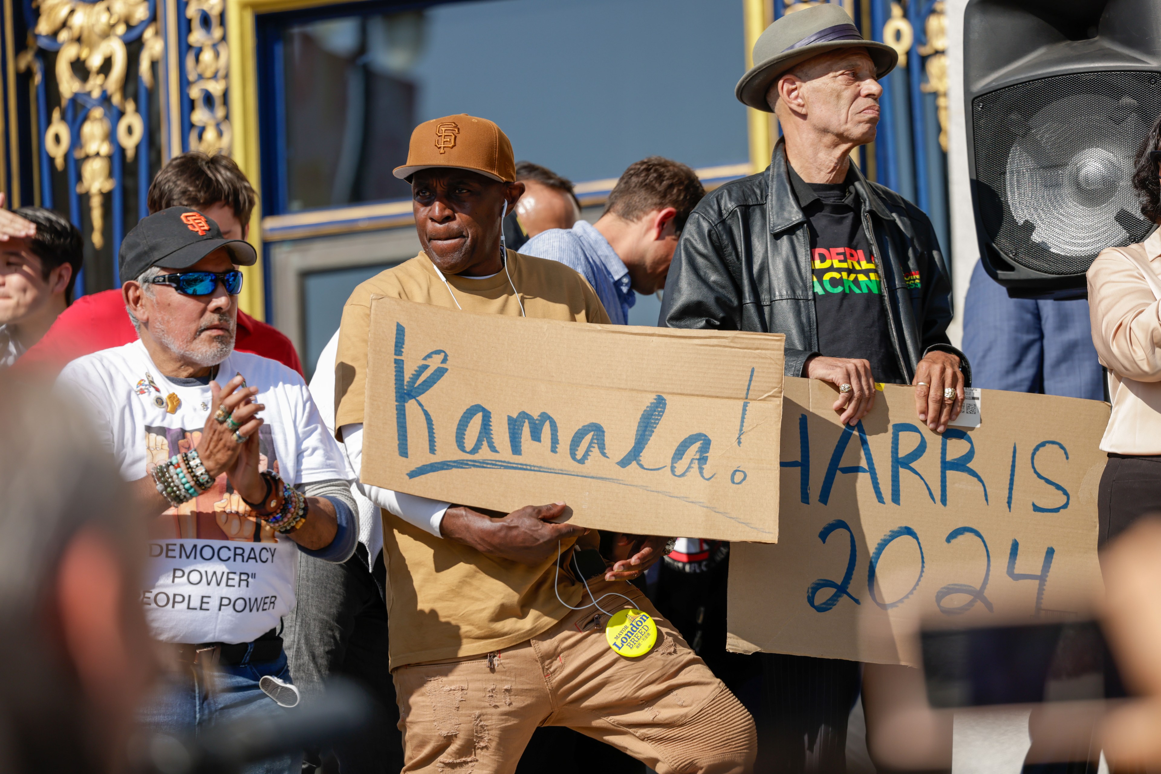 Three men at a rally, holding signs supporting Kamala Harris for the 2024 election, with one reading &quot;Kamala!&quot; and the other &quot;Harris 2024.&quot;