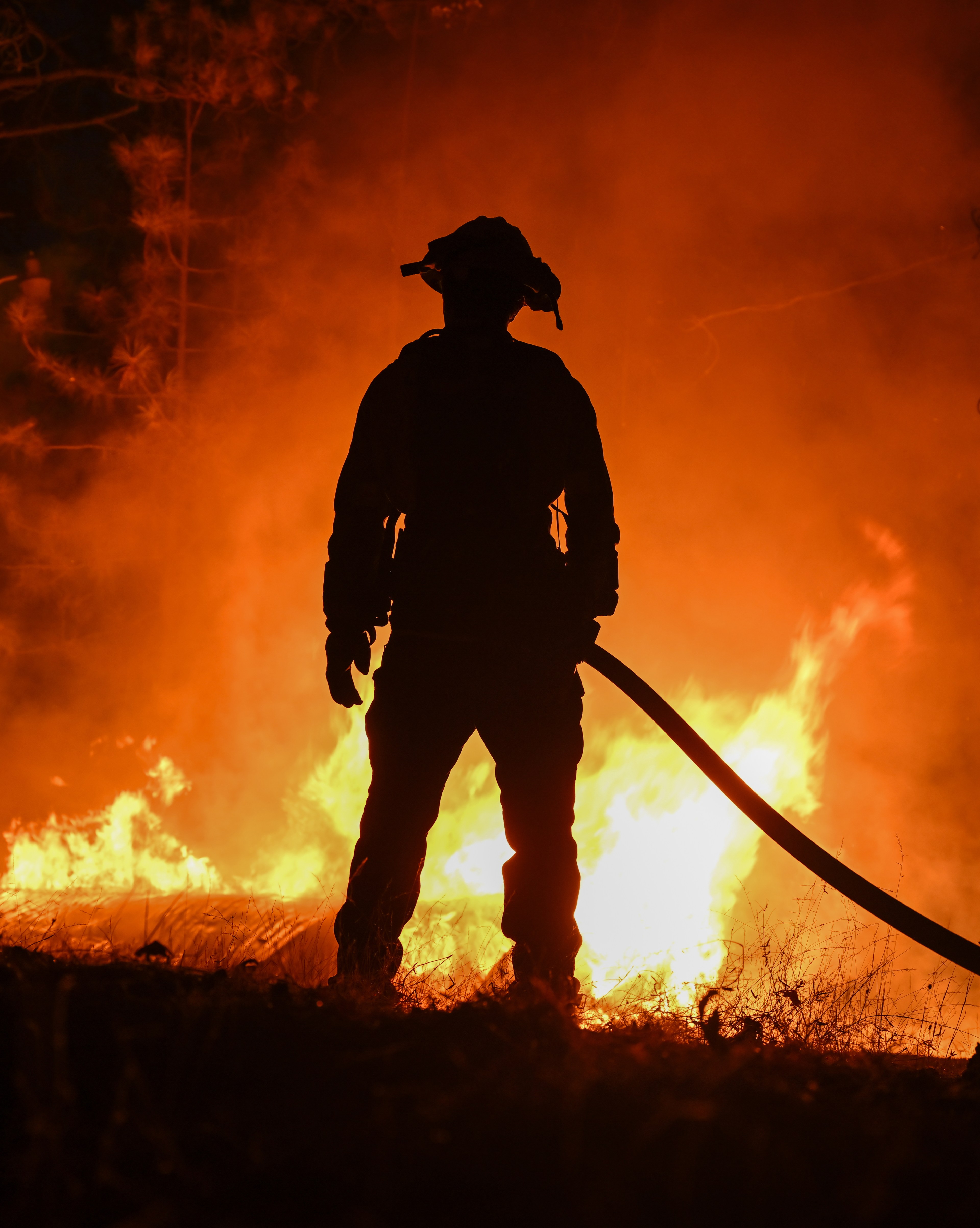 A firefighter in silhouette stands facing a raging fire, holding a hose, with intense flames and smoke illuminating the dark surroundings.