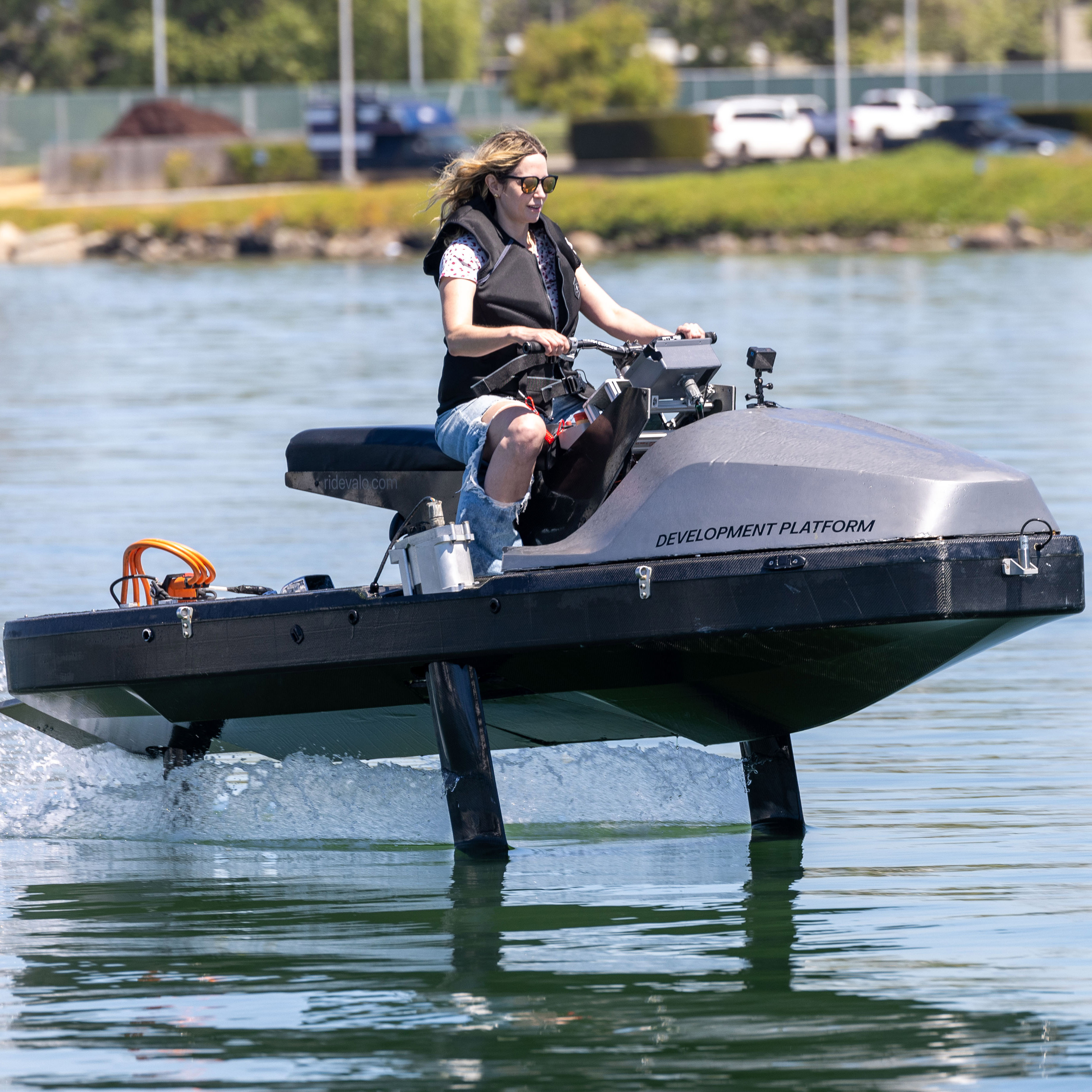 A person in sunglasses and a life jacket is riding a futuristic-looking watercraft labeled "DEVELOPMENT PLATFORM" on a calm body of water.