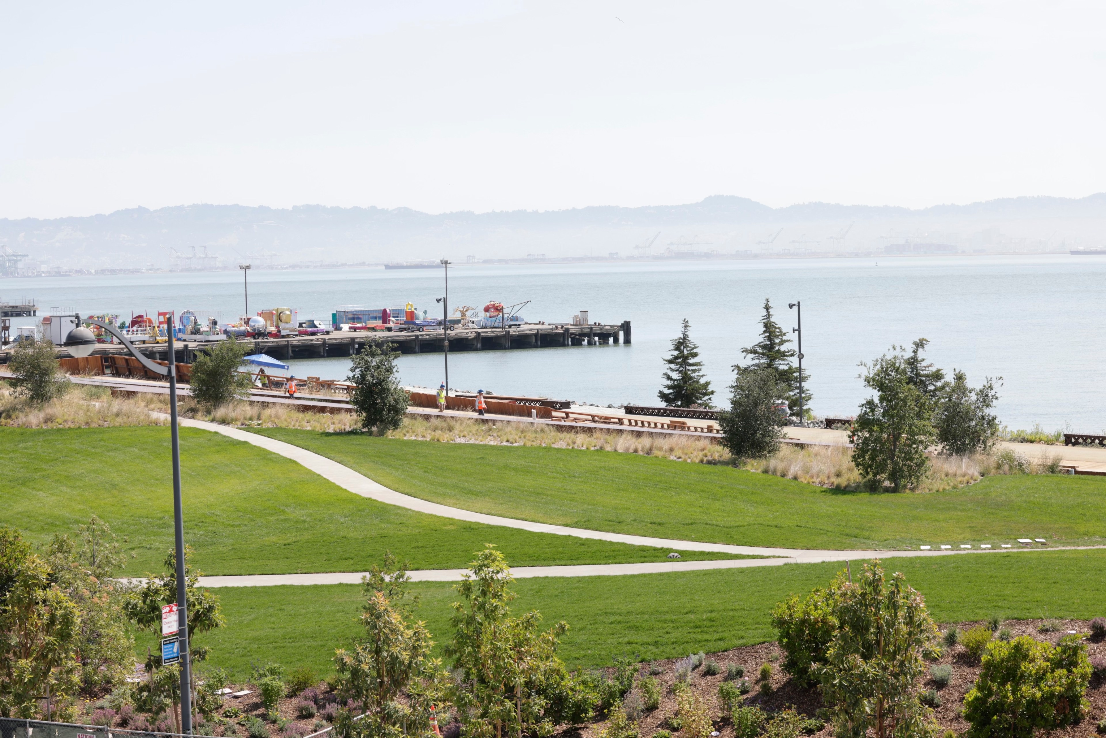 The image shows a waterfront park with a lush green lawn, winding paths, and some trees, leading to a dock with colorful containers and equipment by a calm, expansive body of water.