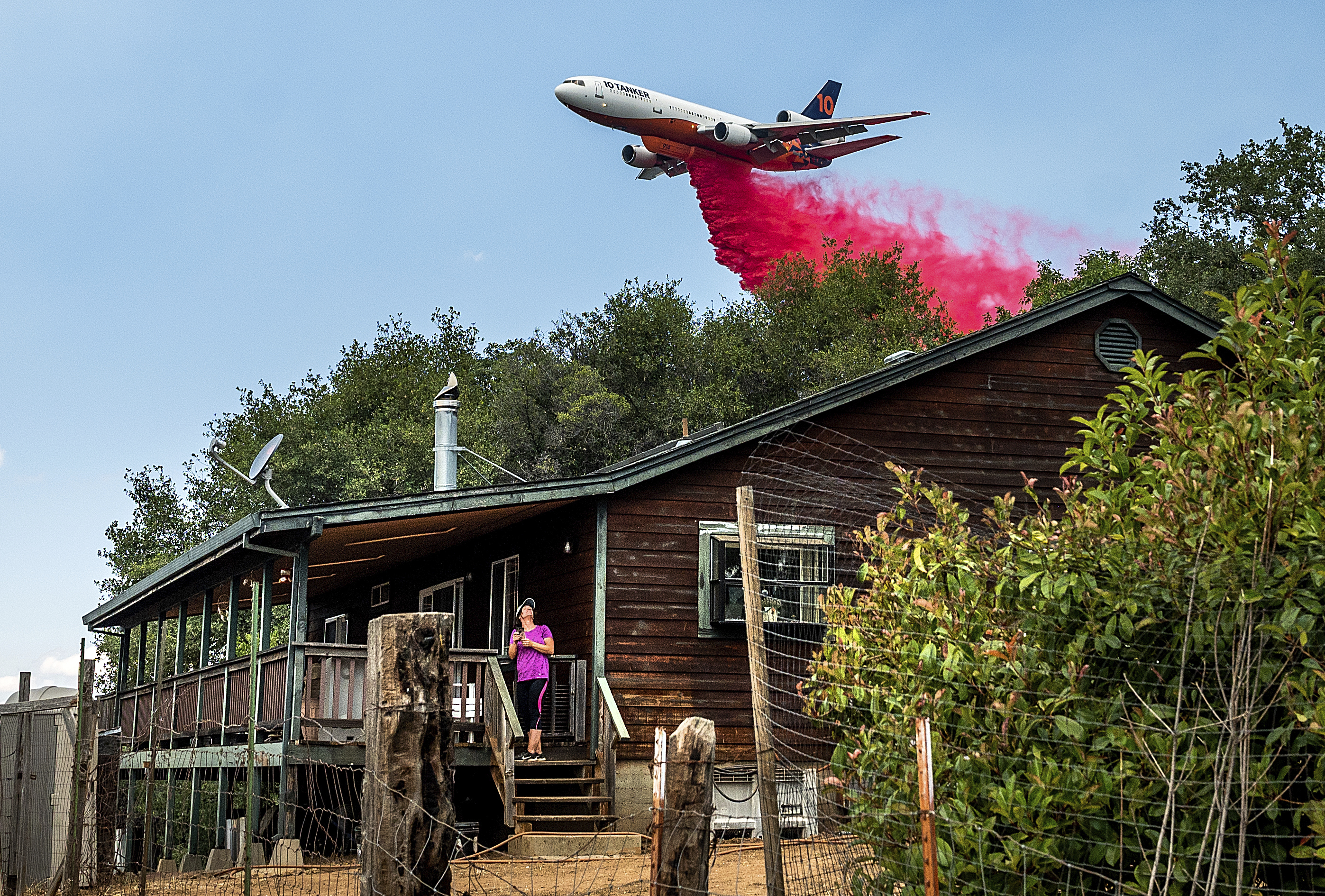 A large air tanker releasing red fire retardant flies low over a cabin in a wooded area, with a person standing on the porch watching.