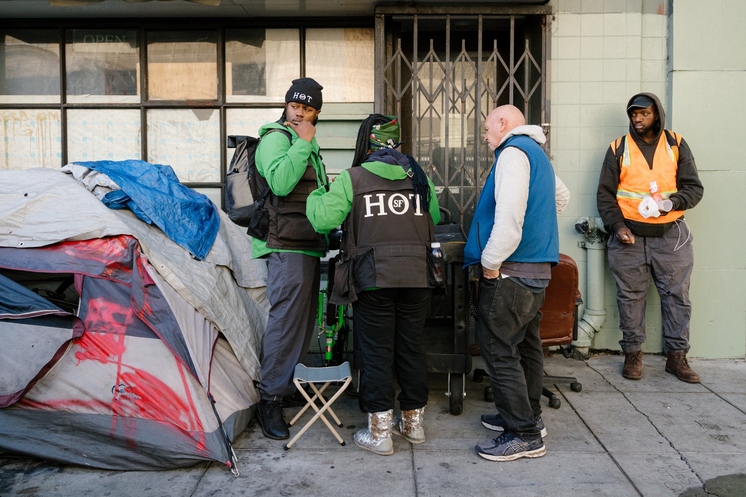 Four people are gathered on a sidewalk near a makeshift shelter. Two wear green and black "HOT" jackets, one wears an orange vest, and another wears a blue vest.