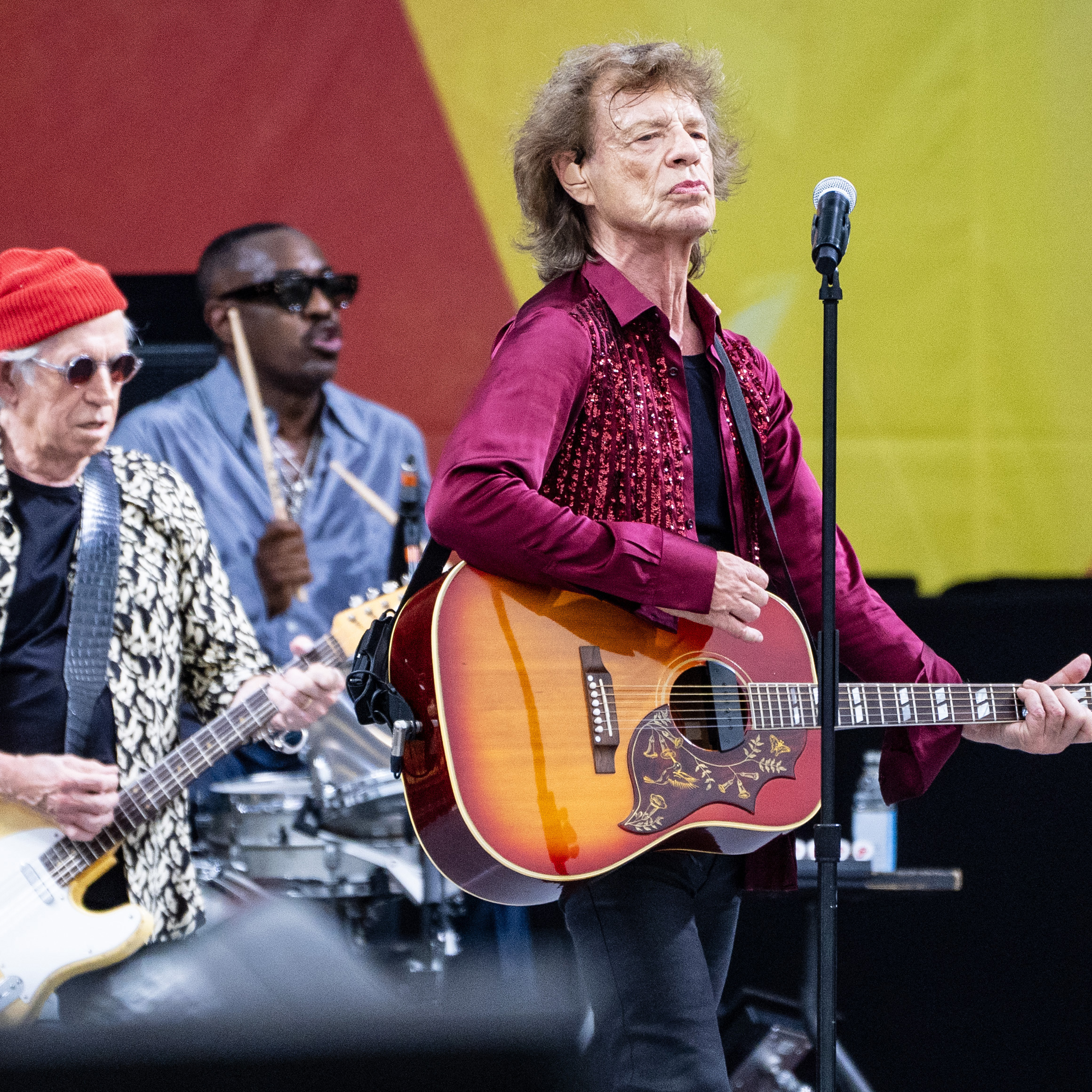 A band is performing on stage. The lead singer, with a guitar and wearing a red shirt, stands at the mic. A guitarist in a red beanie and sunglasses is playing alongside a drummer.