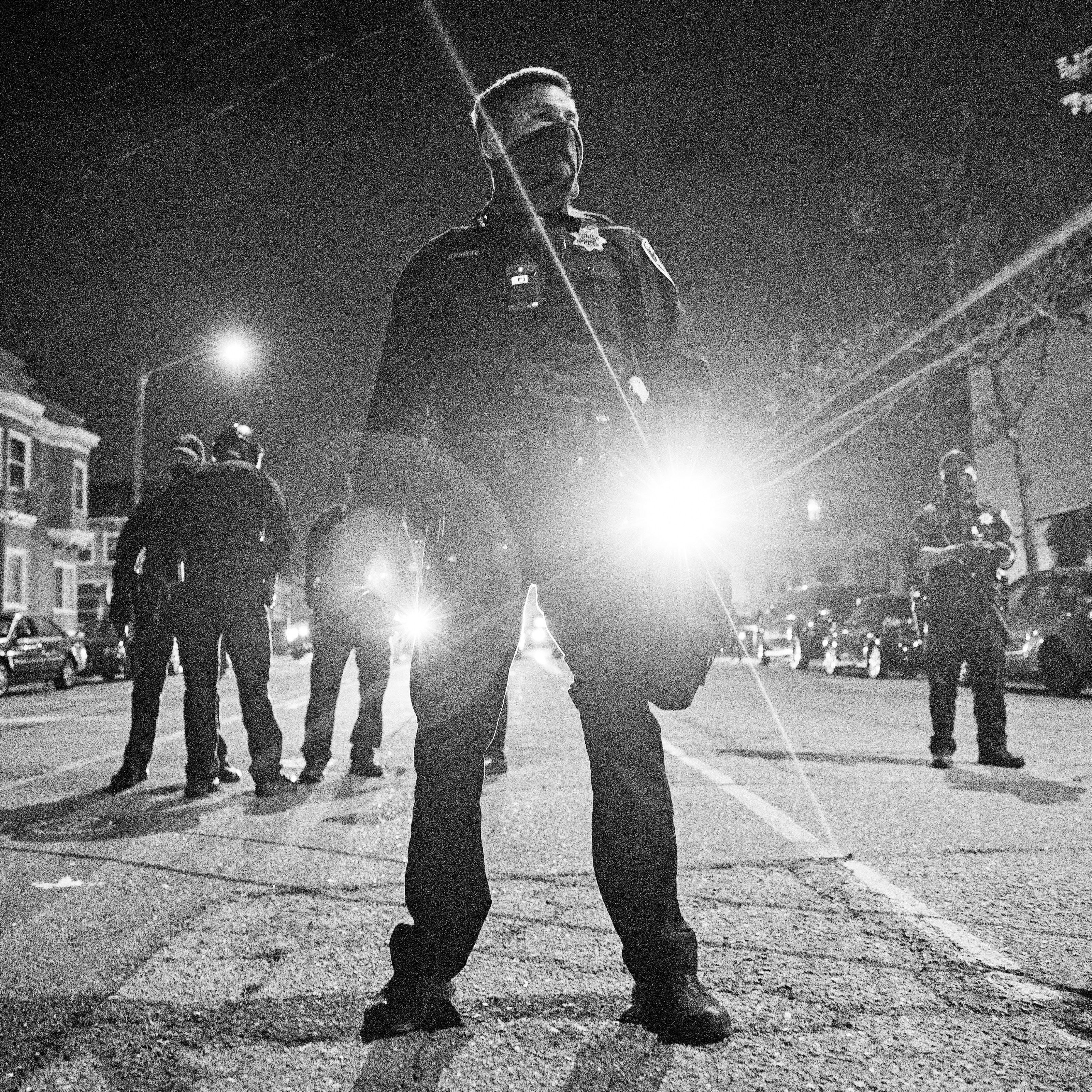 A group of police officers stands in the middle of a street at night, with one officer in the foreground shining a bright flashlight toward the camera.