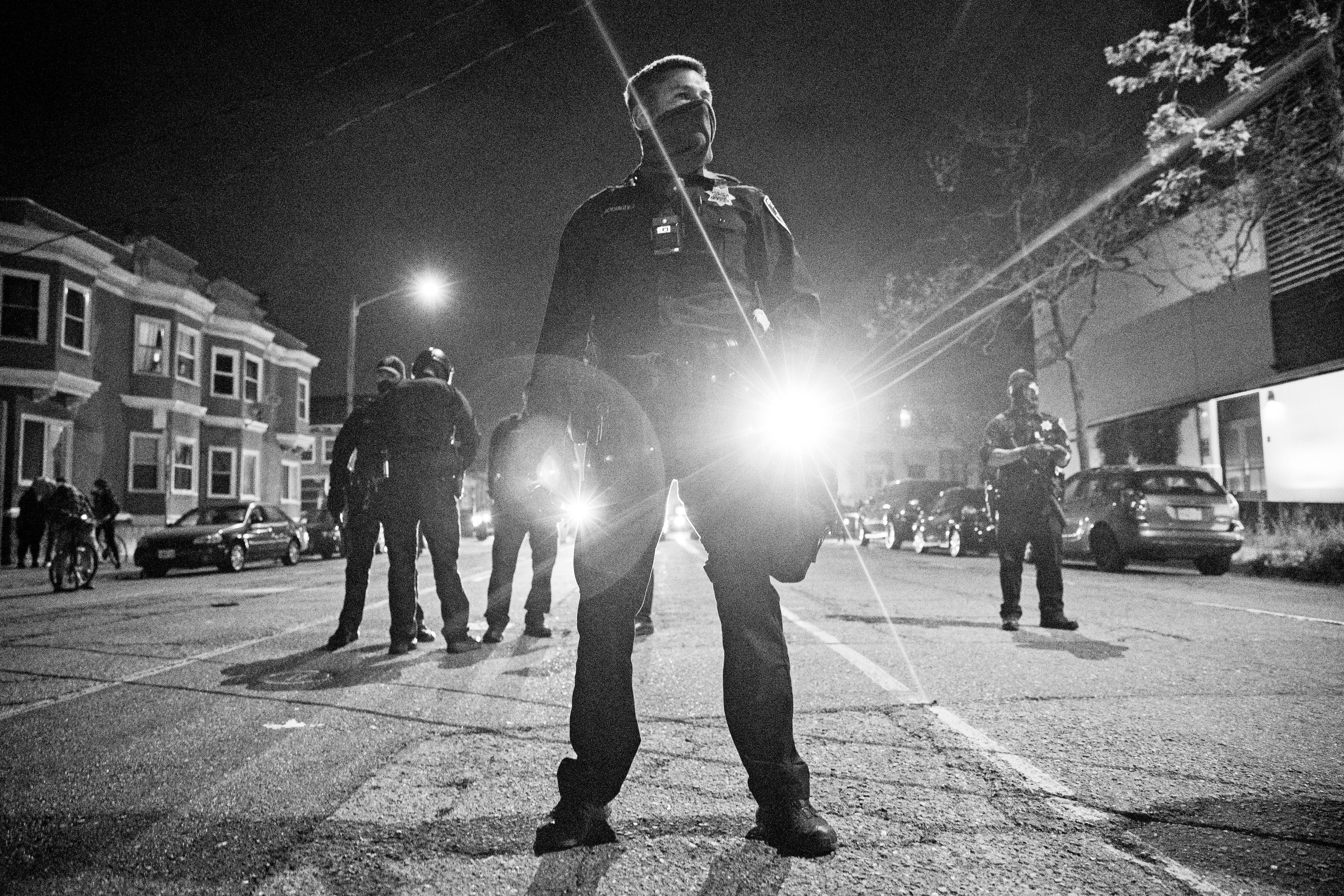 A group of police officers stands in the middle of a street at night, with one officer in the foreground shining a bright flashlight toward the camera.