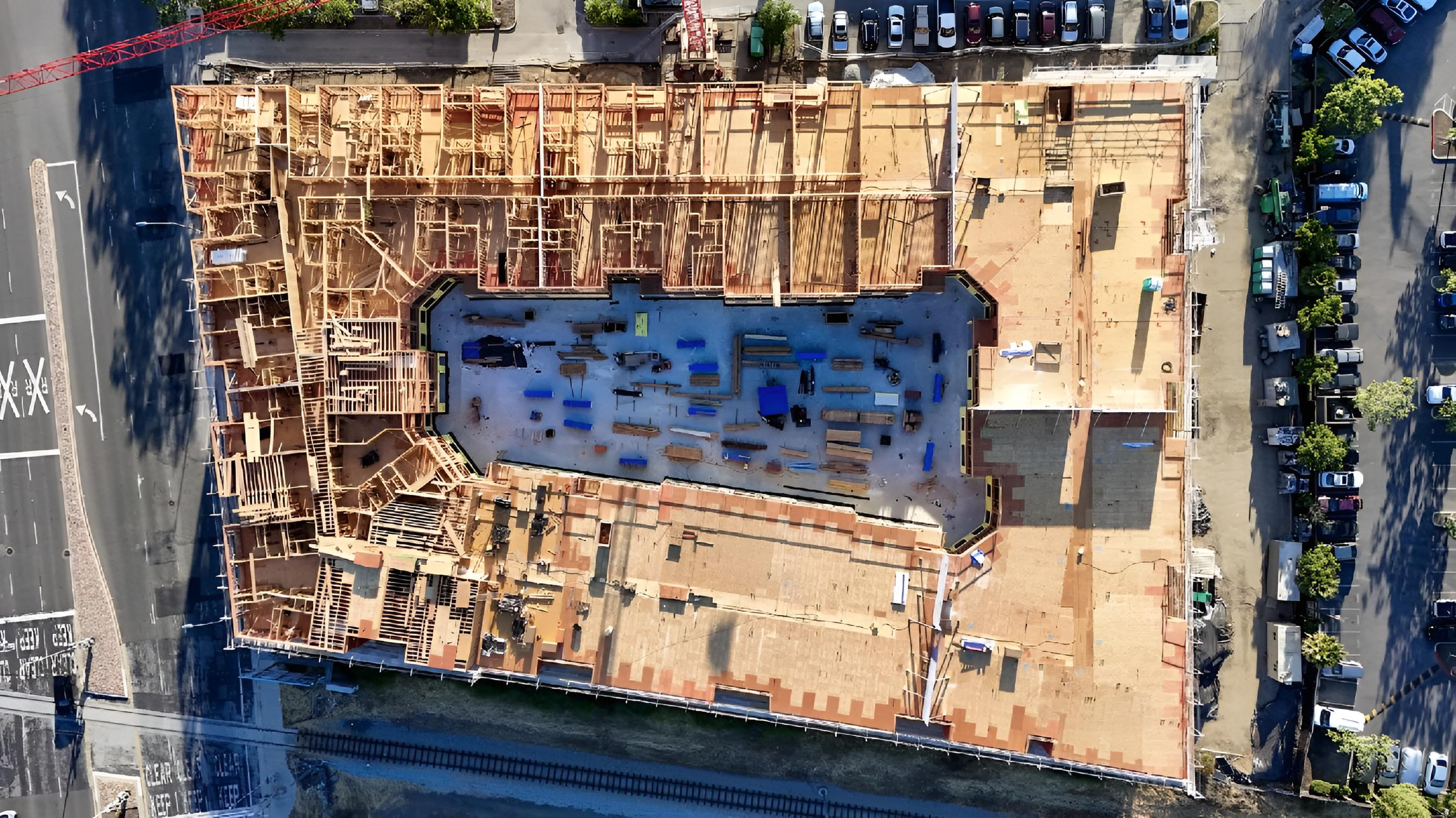 This image shows an aerial view of a large construction site, with the wooden framework of a rectangular building taking shape, surrounded by roads and parking areas.