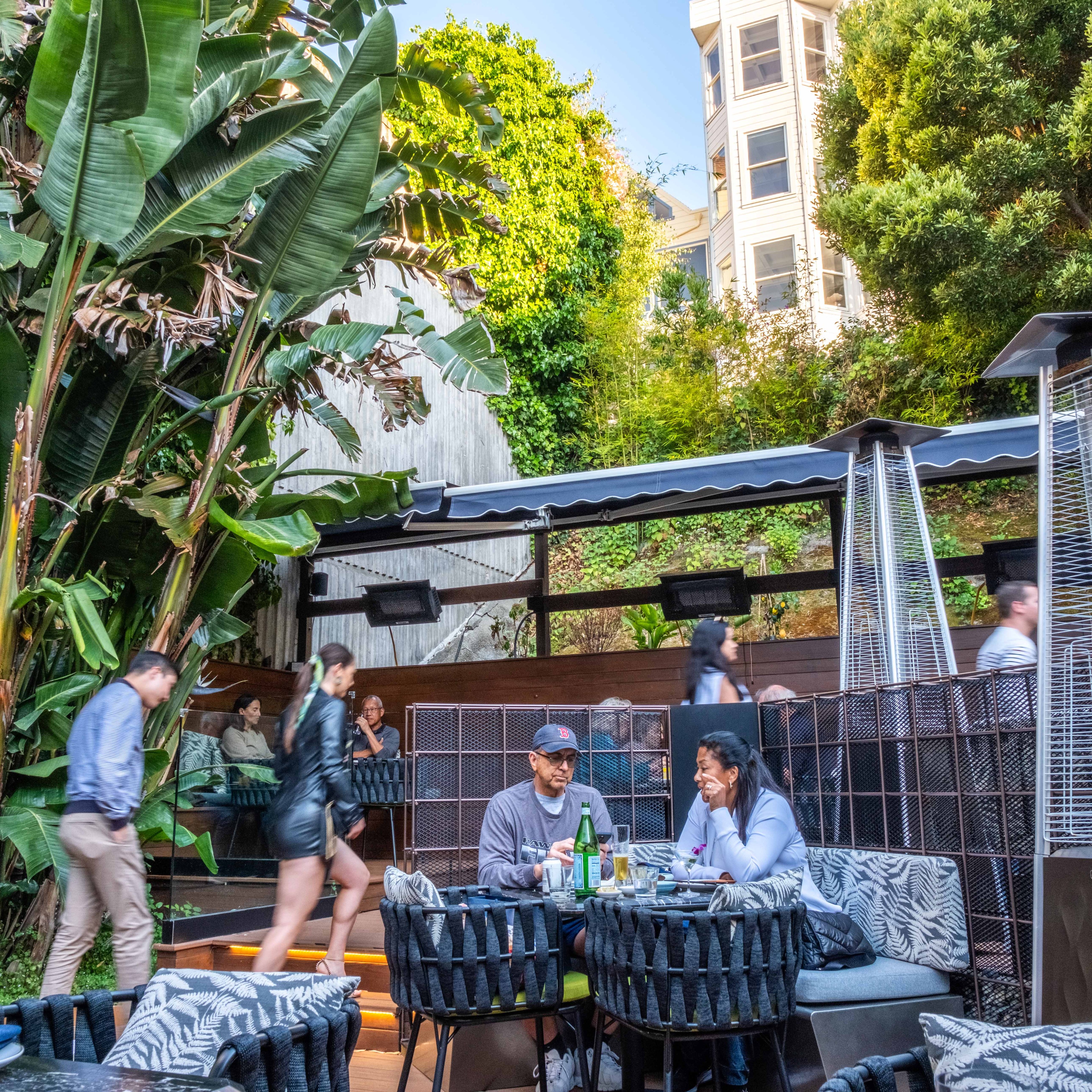 People are dining and walking in a lush outdoor patio with tropical plants and trees, amidst modern furniture and awnings, against a backdrop of hillside greenery.