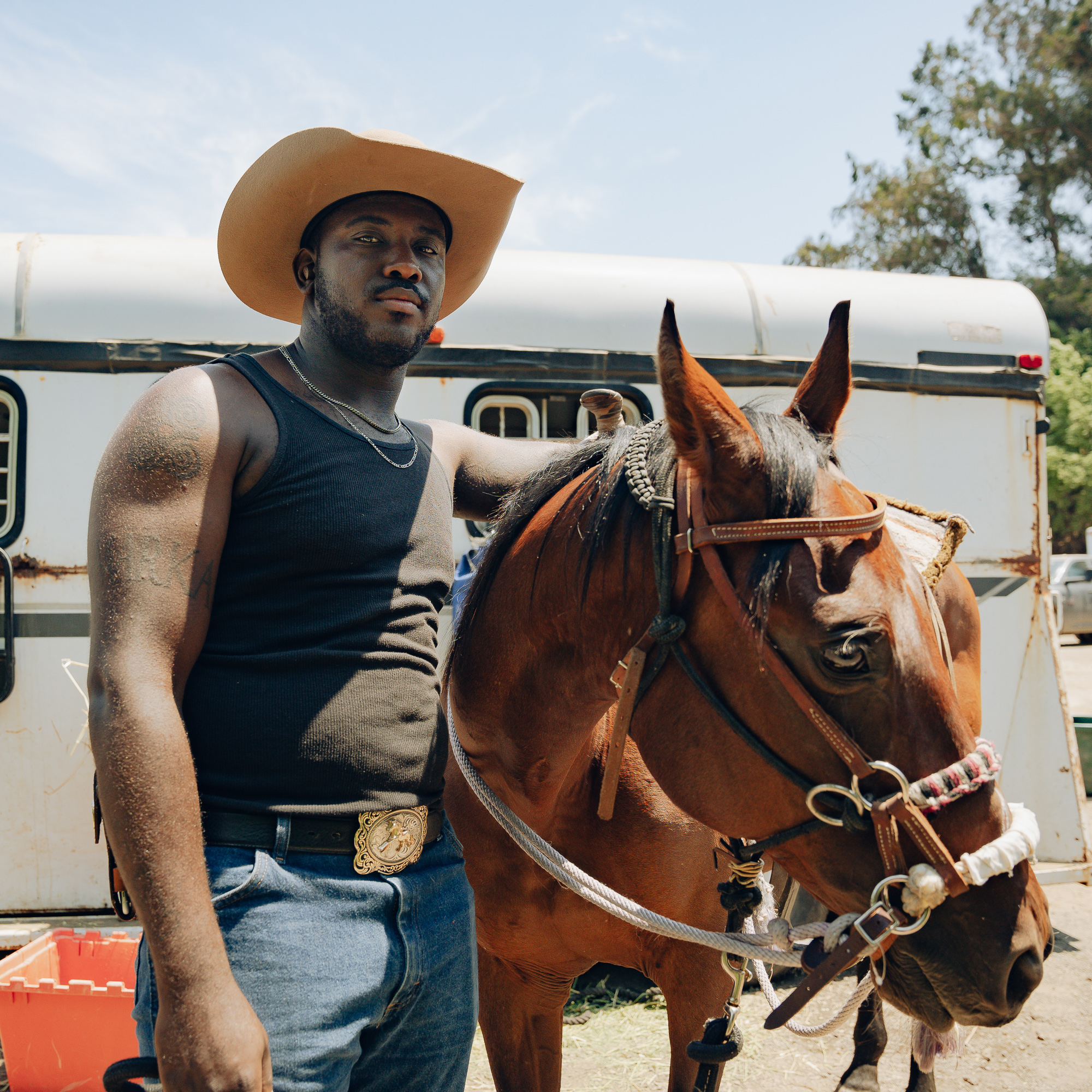 A man in a cowboy hat and sleeveless black shirt stands next to a brown horse, gently holding its reins. A trailer is visible in the background under a clear sky.