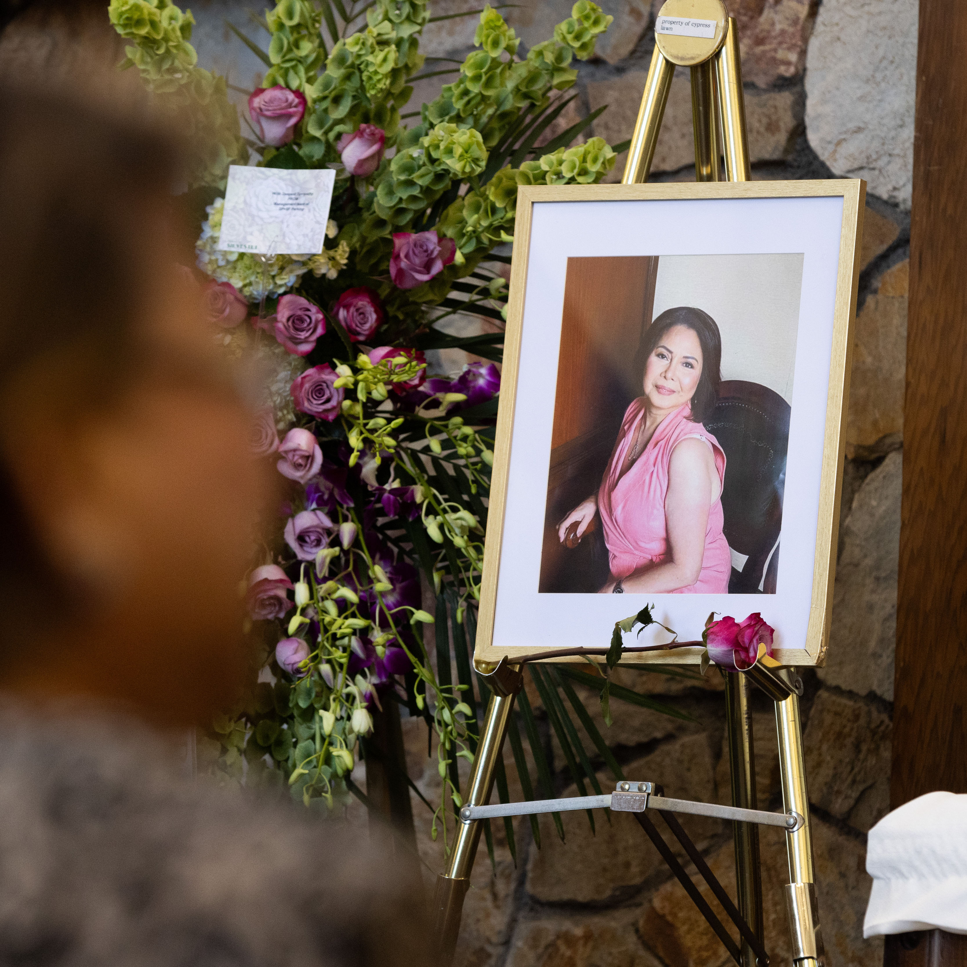 A framed photo of a woman in a pink dress is displayed on an easel next to a bouquet of purple and green flowers. A person is blurred in the foreground.