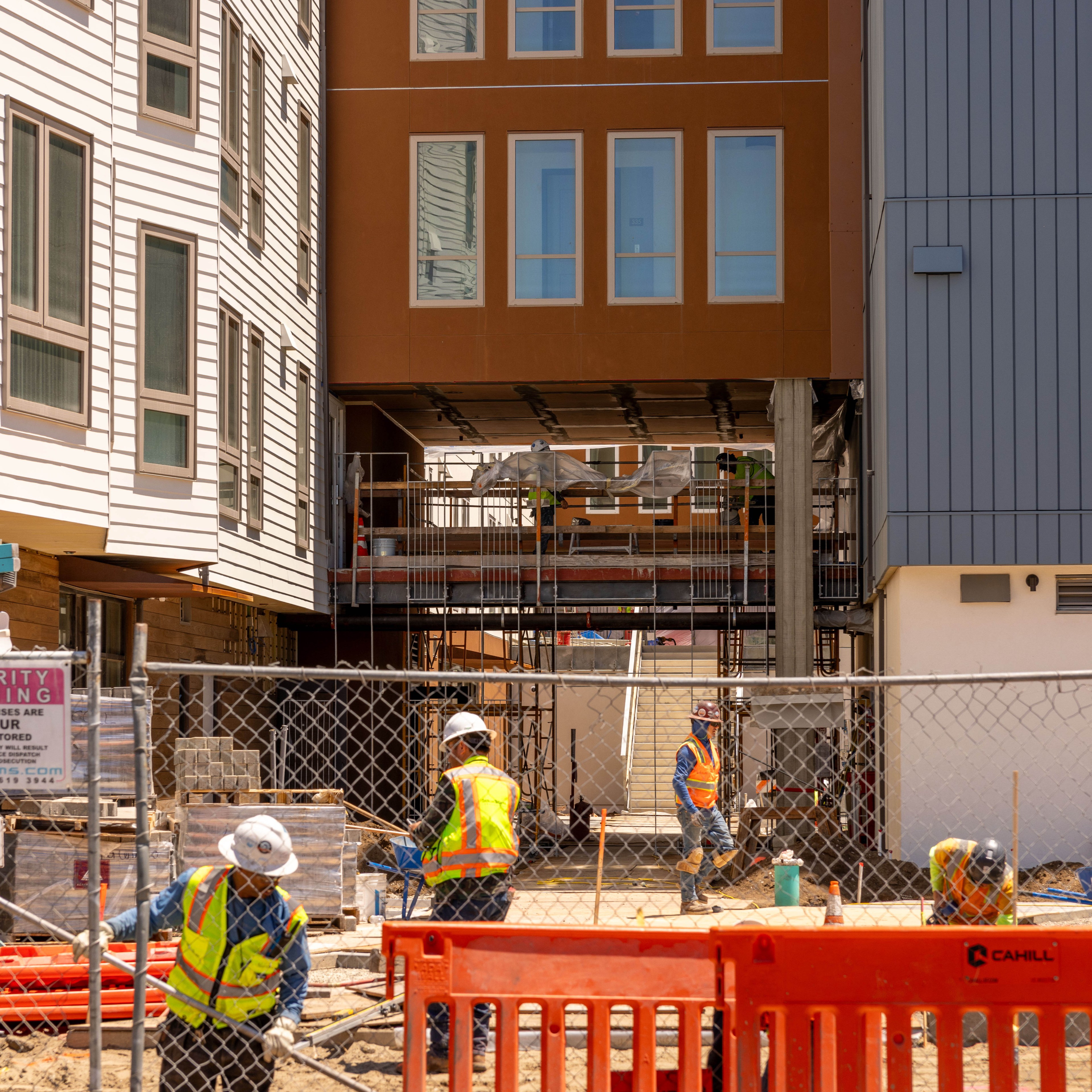 Construction workers in safety vests and helmets work on a building site between three multi-story buildings, one white, one brown, and one gray, behind a chain-link fence.