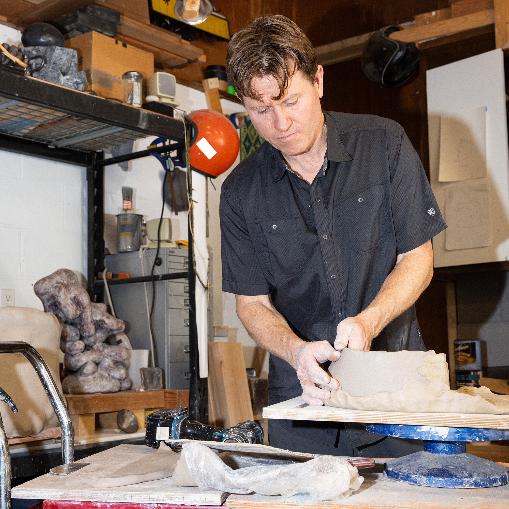 A man in a workshop is sculpting clay on a table, surrounded by various tools and materials. He appears focused on his work, with shelves and equipment in the background.