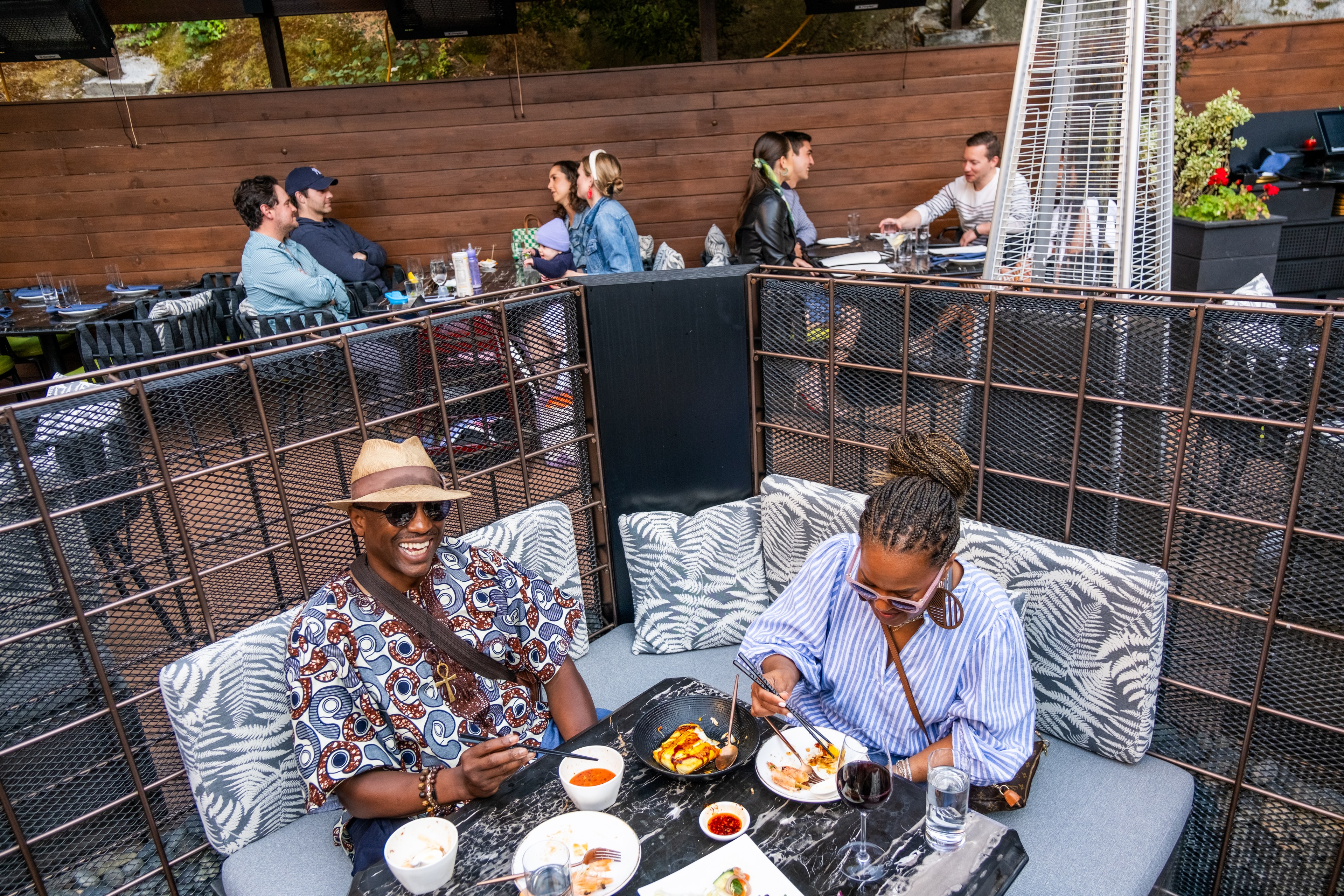 Two people are eating and laughing at an outdoor restaurant, with six more people dining at separate tables in the background. The setting appears relaxed and lively.