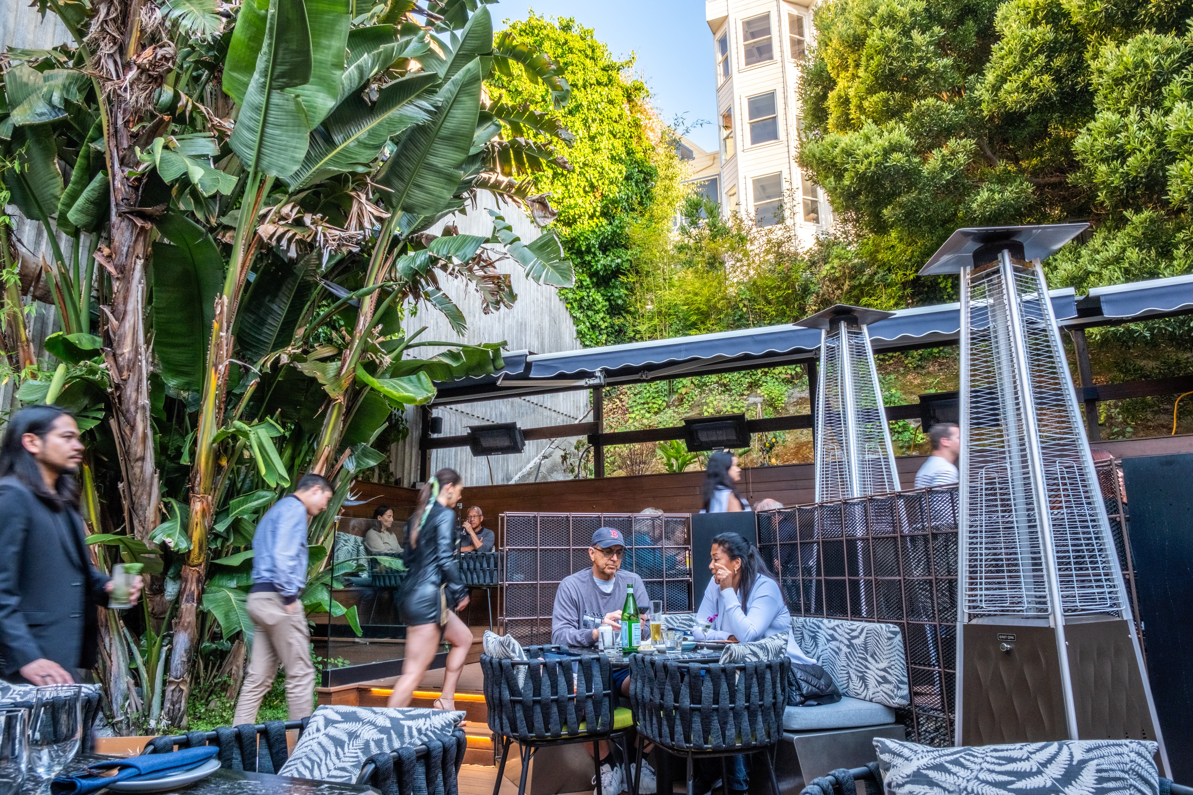 An outdoor restaurant setting with tropical plants, people sitting and eating at tables, and heaters around. Some patrons walk by while others sit and converse.