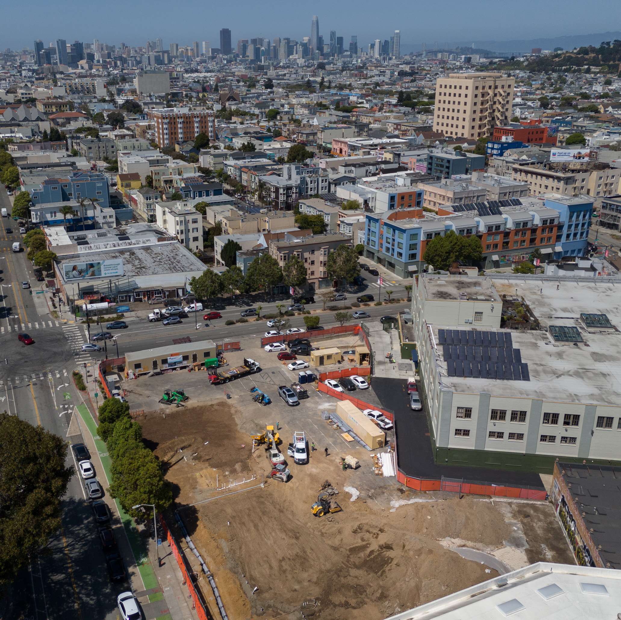 The image shows an urban construction site with machinery and workers, surrounded by streets, cars, and buildings, with a city skyline visible in the distance.