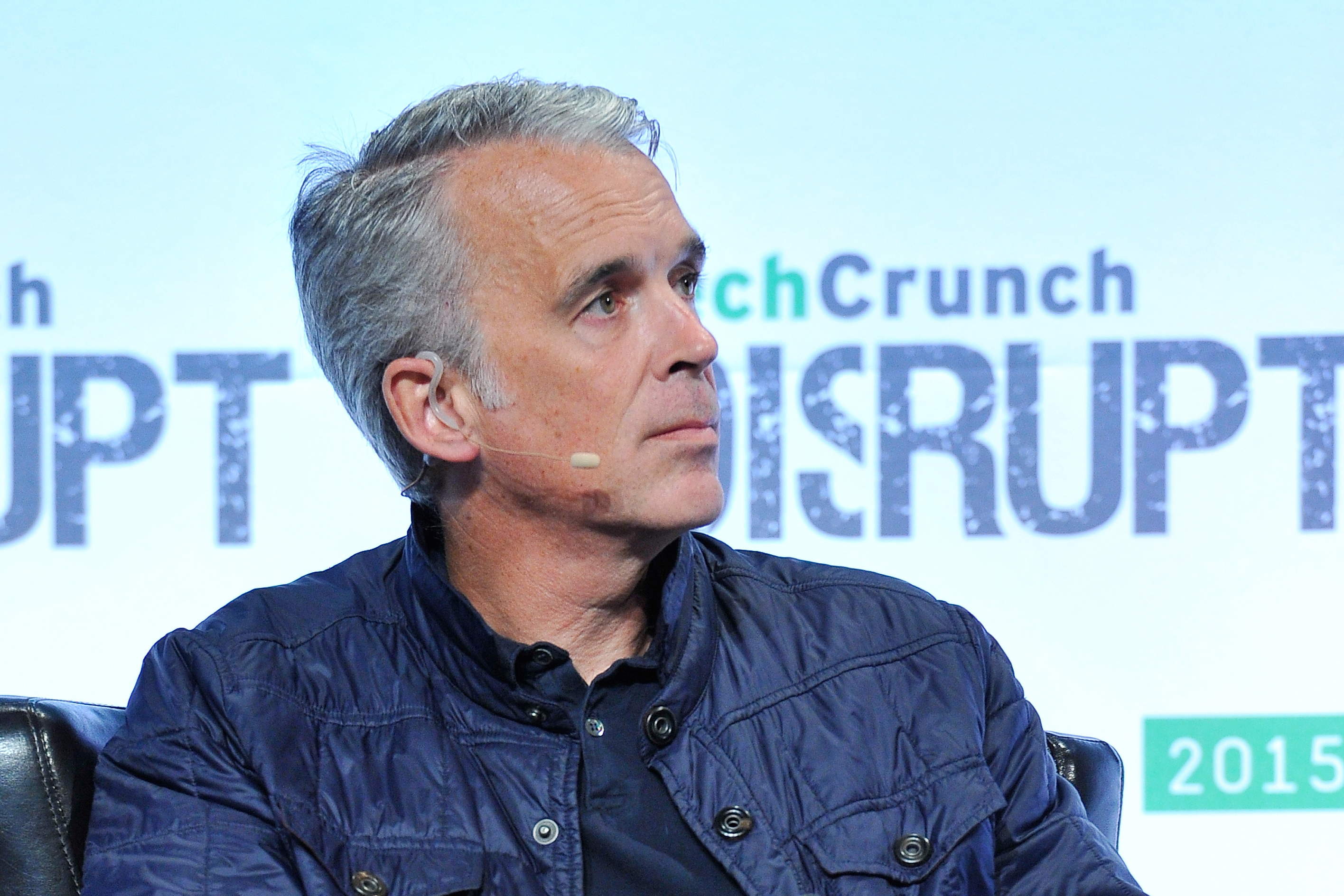 A man with short gray hair sits on a black chair, holding a tablet. He wears a dark blue jacket, jeans, and brown loafers. The background reads "TechCrunch Disrupt 2015."