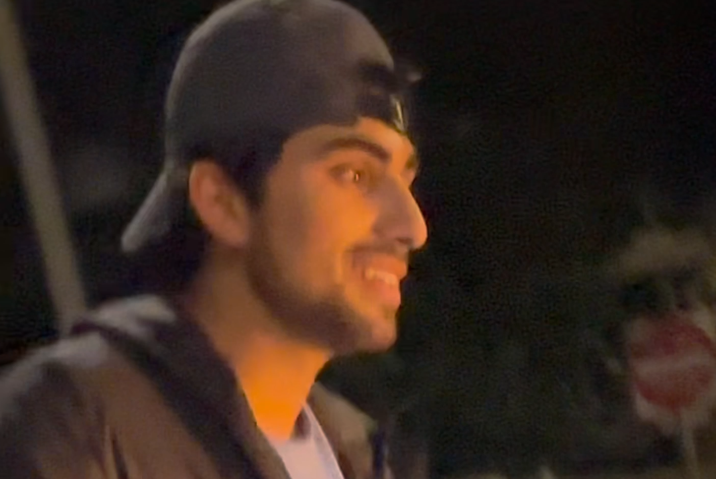 A man wearing a backwards baseball cap, hooded sweatshirt and jacket smiles in low light at nighttime.