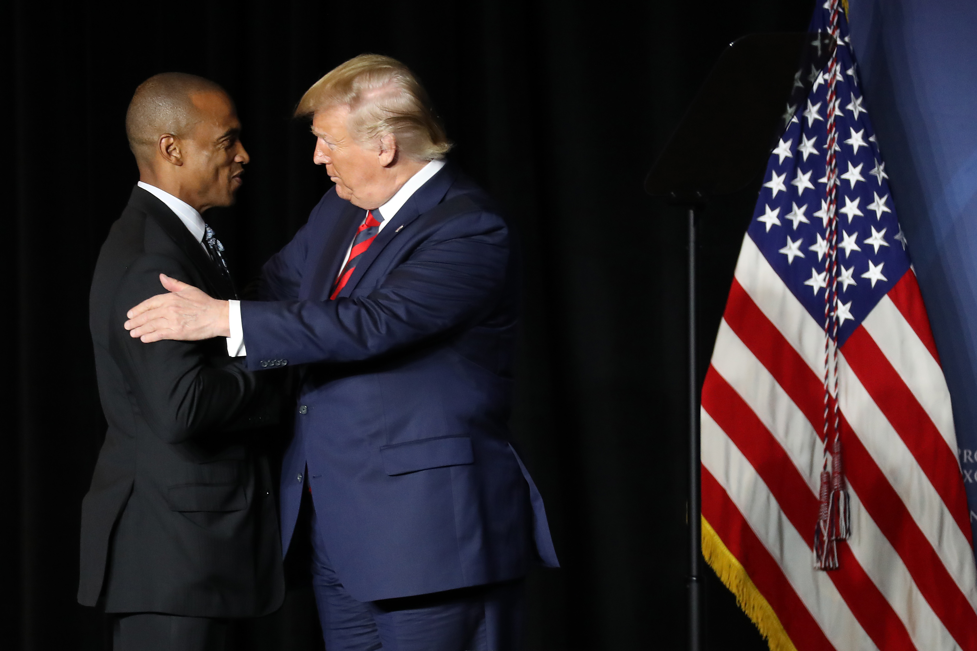 Two men in suits are shaking hands on a stage with a U.S. flag beside them, one in a blue suit and red tie, and the other in a black suit.