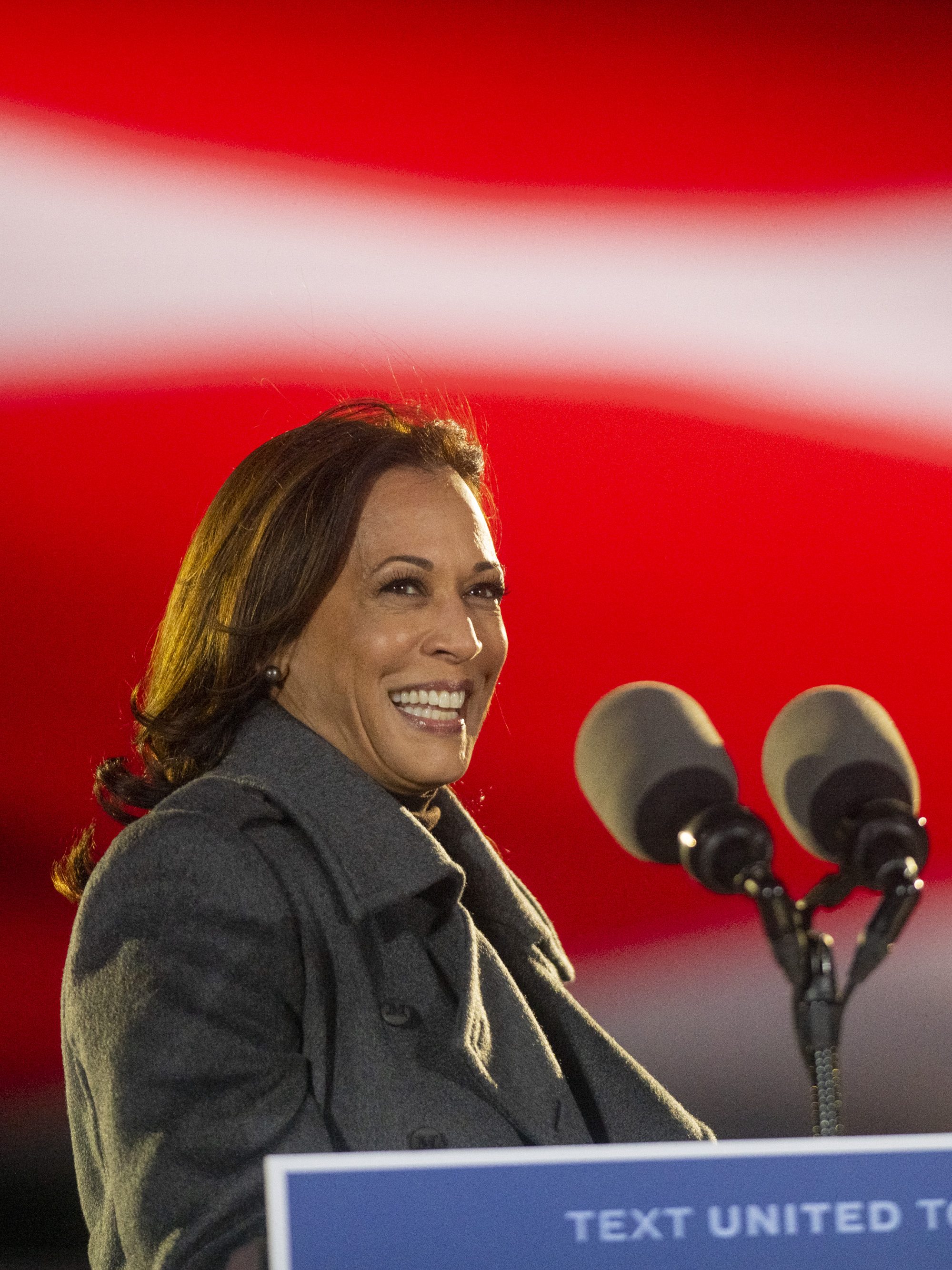 A woman in a gray coat is smiling behind a podium with two microphones. The background features a blurred, waving flag pattern in red, white, and blue.