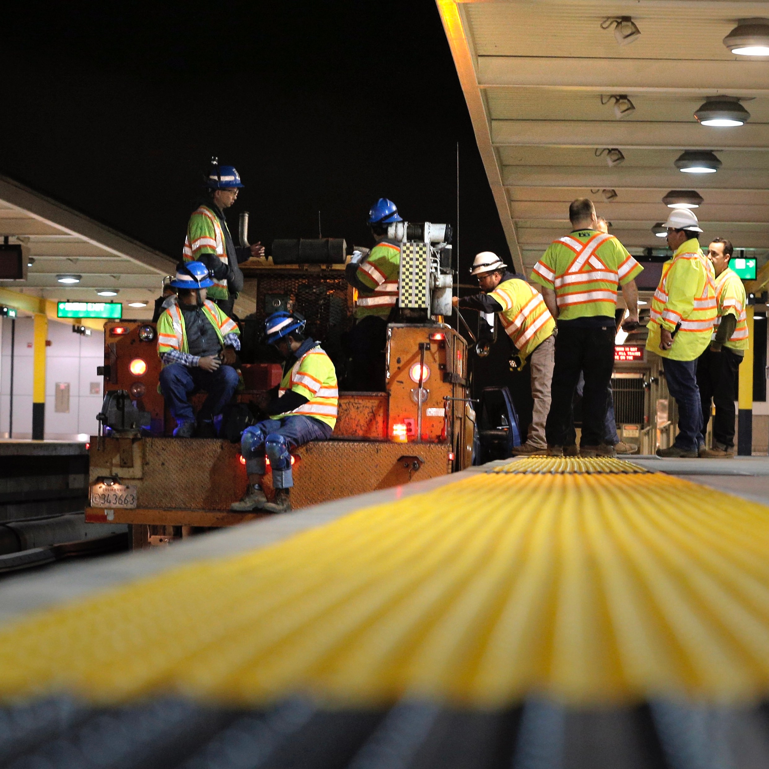 The image shows a group of construction workers in reflective vests and hard hats, both standing and seated, on a subway platform at night, inspecting equipment.