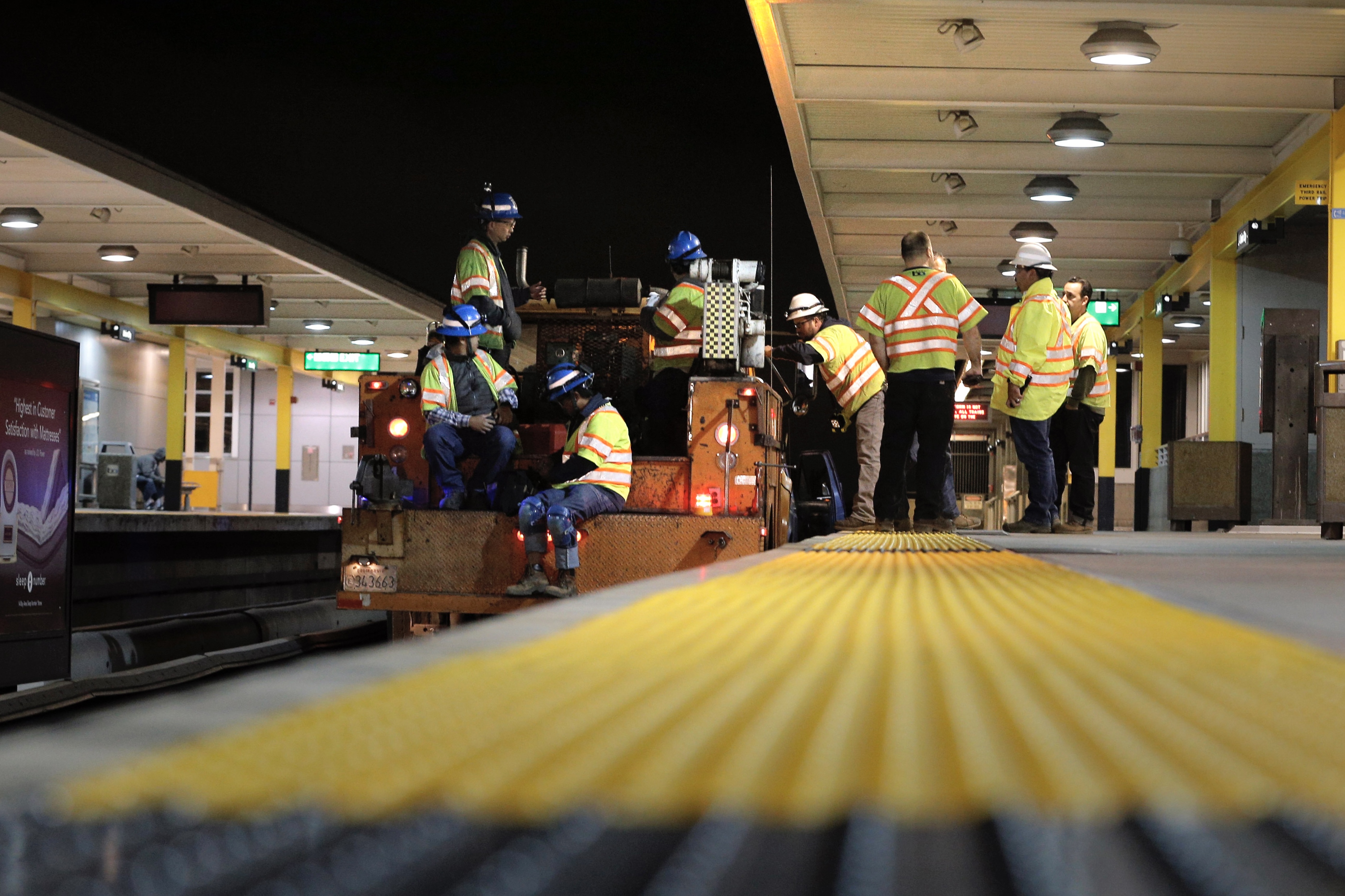 The image shows a group of construction workers in reflective vests and hard hats, both standing and seated, on a subway platform at night, inspecting equipment.