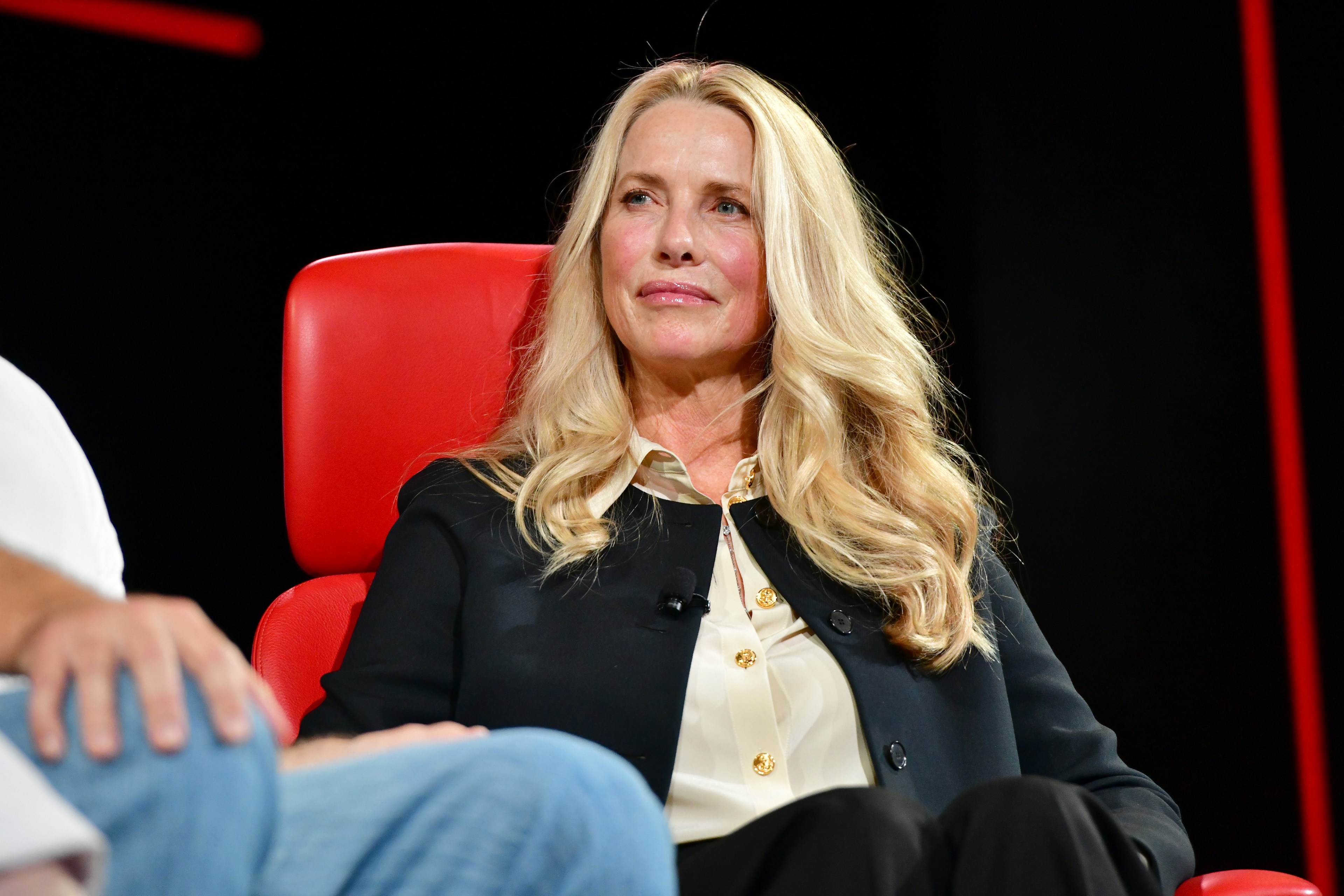 A woman with long blonde hair is sitting in a red chair, wearing a white blouse and a dark blazer, looking slightly to the side with a calm expression.