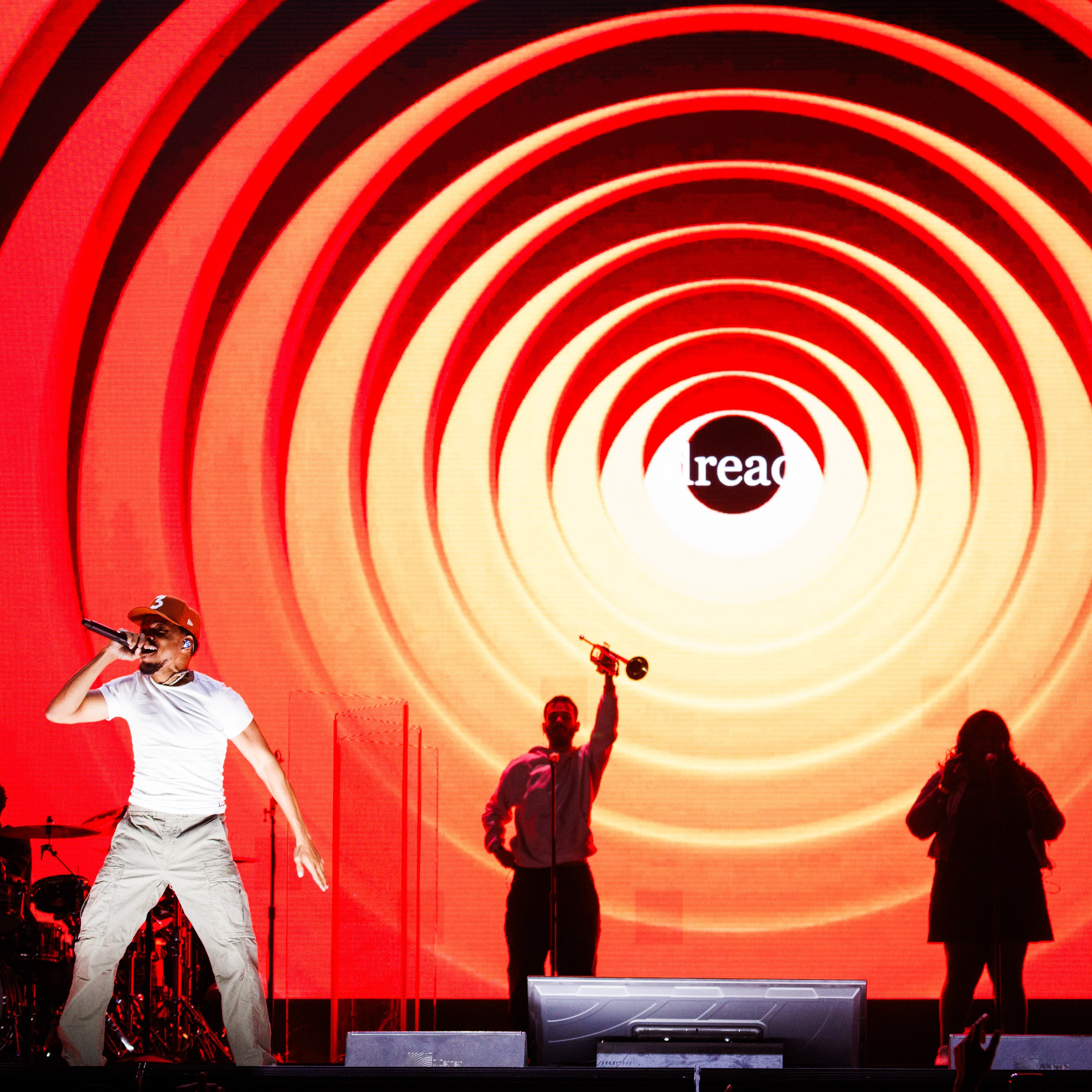 A performer is energetically singing on stage with a red and orange spiral backdrop. Silhouetted band members and a "read" text in the background are also visible.